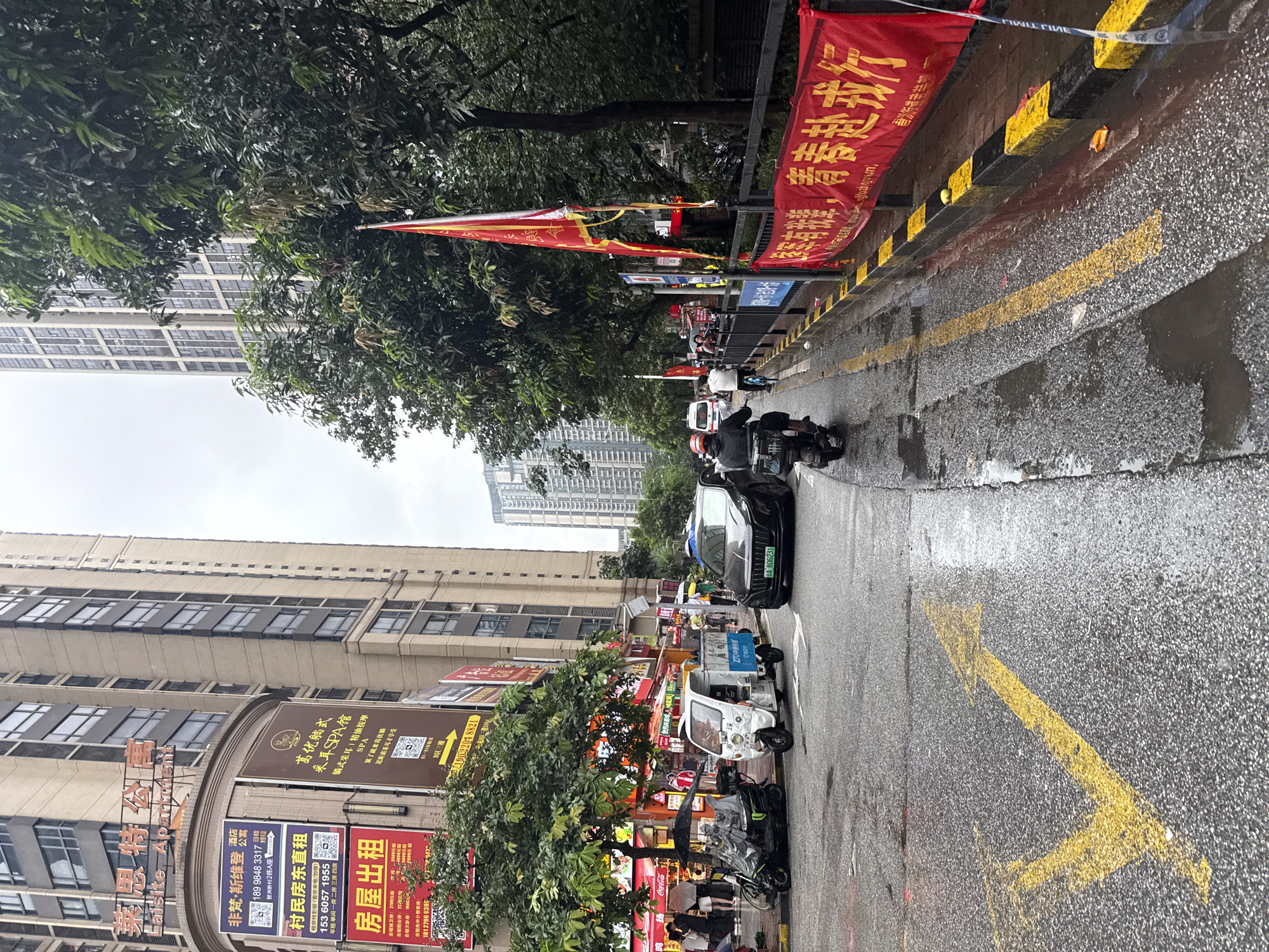 This is a street scene in Haizhu District, China, on a rainy day. The road is wet and reflective. In the foreground, a yellow arrow is painted on the asphalt, indicating a turn. To the left, a black scooter with a rider wearing a helmet and a backpack is turning. Next to it is a white, three-wheeled vehicle, likely a small delivery truck. Further down the street, a black car is driving, its license plate visible as "WA 806231".
On the right side of the street, a red banner with yellow Chinese characters is hung along a black fence. The banner appears to be a promotional or celebratory message. Behind the fence are trees with dense foliage, and further in the background, several tall, modern buildings are visible. The sky is overcast, contributing to the dim lighting of the scene. People are present, some holding umbrellas, suggesting ongoing pedestrian activity despite the weather. Signs on the buildings indicate commercial establishments, including what appears to be an apartment complex and businesses.