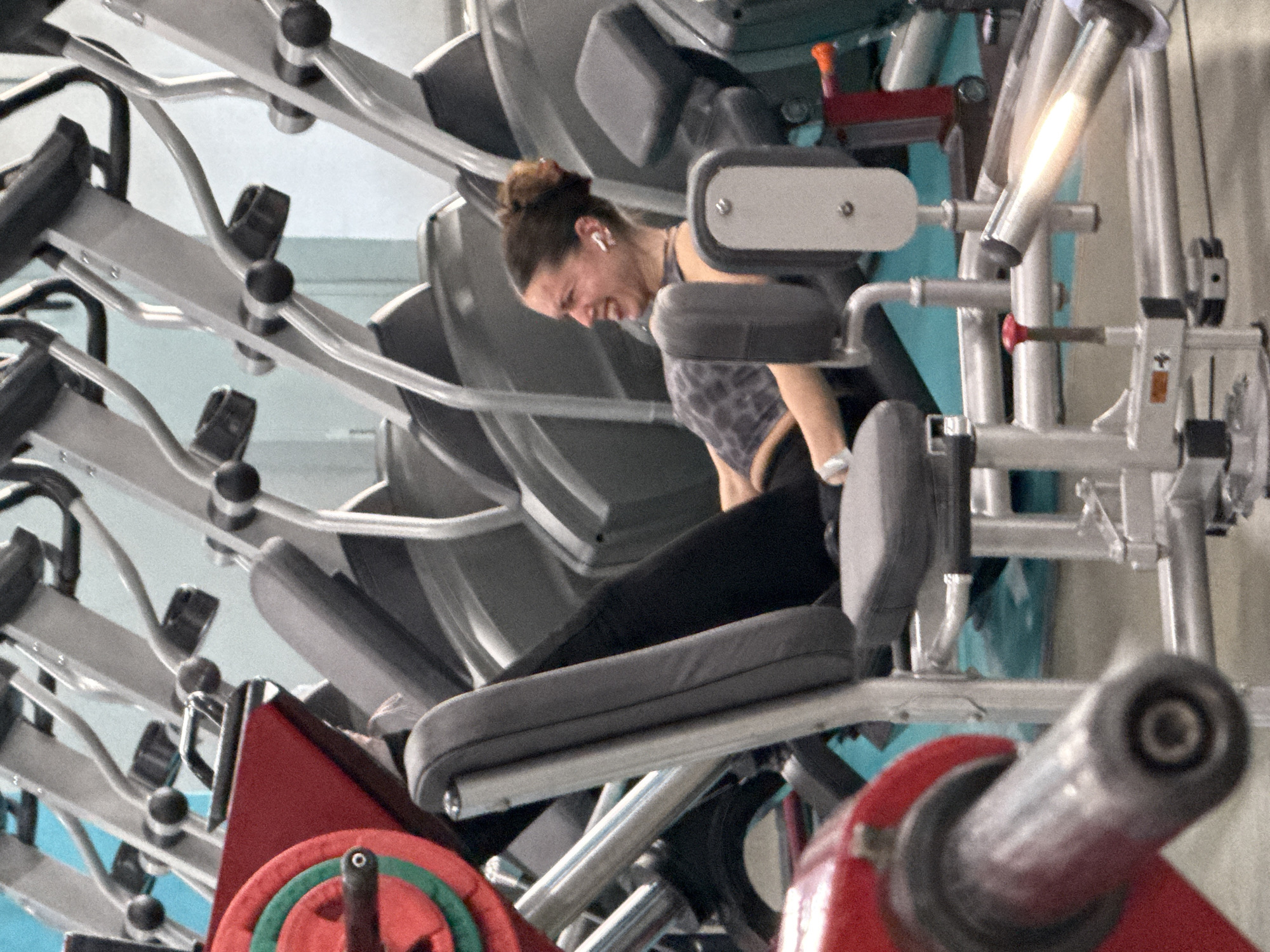 A woman with her hair in a bun and wearing a gray and black patterned sports bra and black leggings is using a weight machine. She has a grimace on her face, suggesting she is exerting herself. The machine is red, gray, and silver, with various levers, pads, and weights visible. The background is out of focus but appears to be a gym setting with other machines. The location in the city of Yekaterinburg, Russia is not discernible from the image.