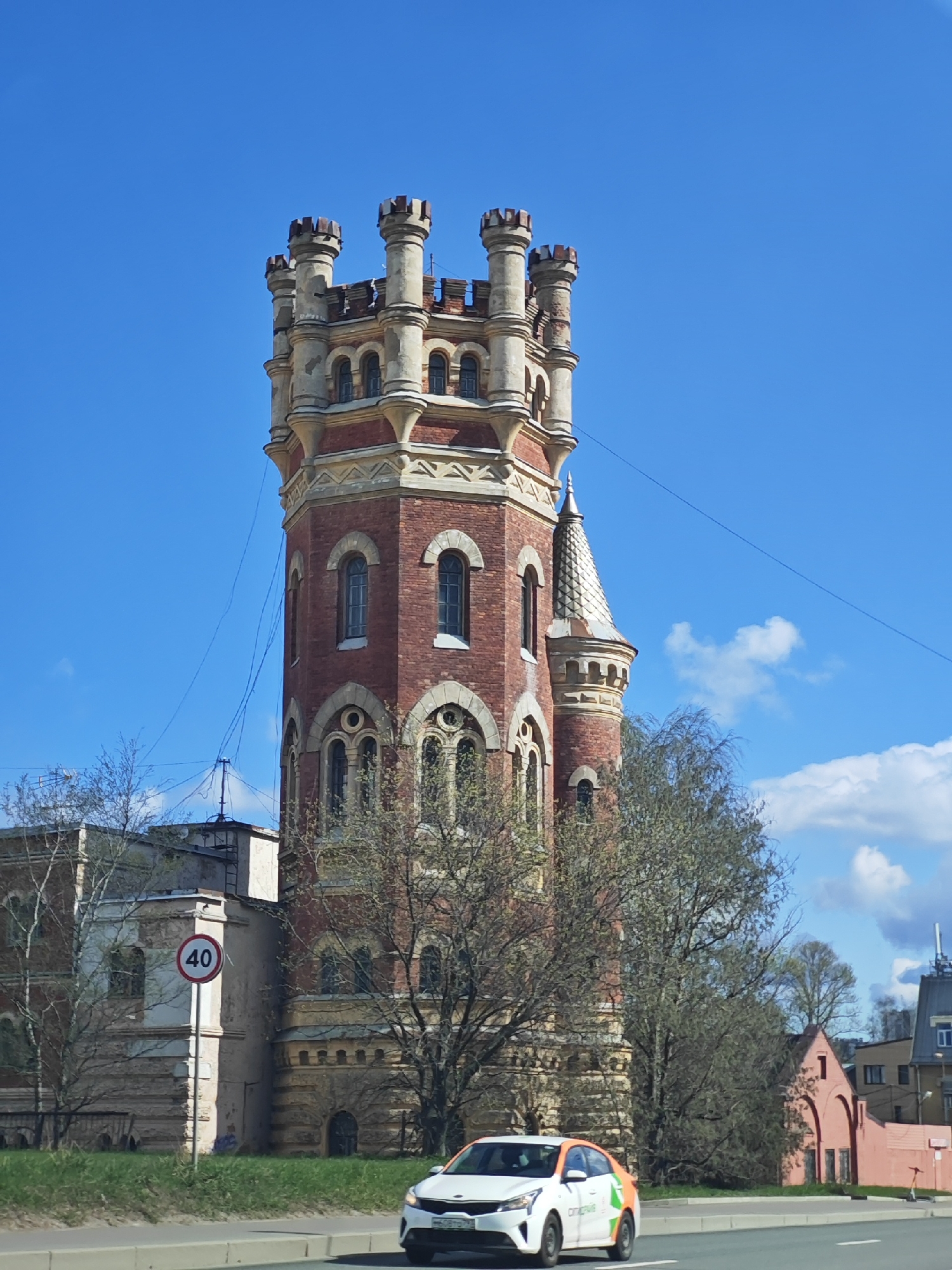The image captures the Water Tower of the former Peterhof Road Water Supply System in Saint Petersburg, Russia. The tower, constructed of red brick with decorative stone elements, stands prominently against a clear blue sky. Its architecture features multiple levels, arched windows, and a series of crenellated turrets at the top. Power lines traverse the upper part of the structure.
In the foreground, a white car with a green and orange side stripe is visible on the road. A speed limit sign (40 km/h) is placed along the roadside, in front of a section of the adjacent building. Deciduous trees with budding leaves partially obscure the view of the tower and surrounding structures. Other buildings, including a pink building, are visible in the background.