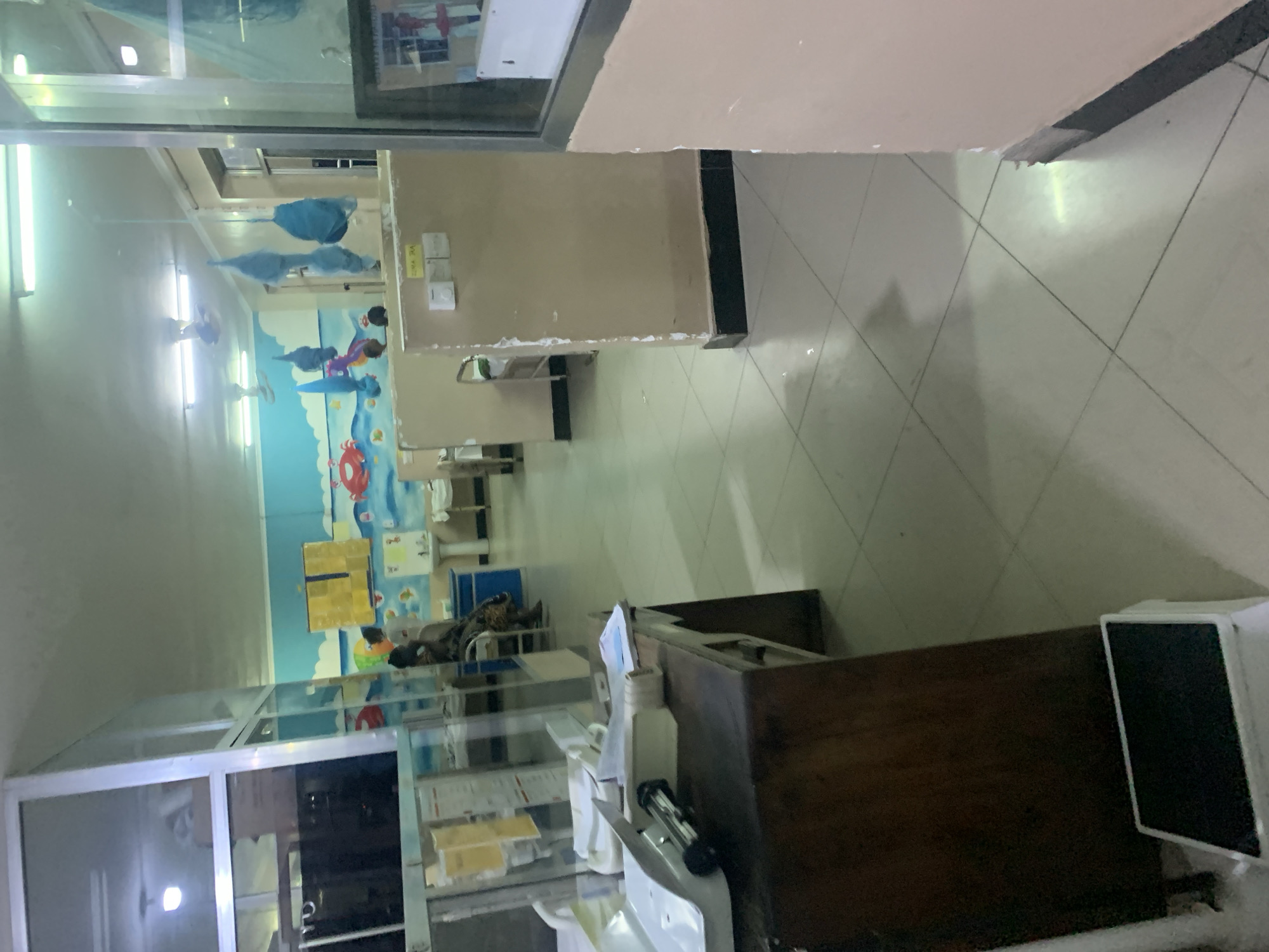 The image depicts the interior of a hospital in Dar es-Salaam, Tanzania. The scene includes a hallway with a children's mural on the wall, featuring ocean-themed artwork. A reception desk and display cabinets are visible, with various medical supplies and equipment on the counter. A person lies prone on a hospital bed in the background. The floor is tiled, and the walls are painted beige. Fluorescent lights illuminate the space. The presence of Tanzanian currency implies the location.