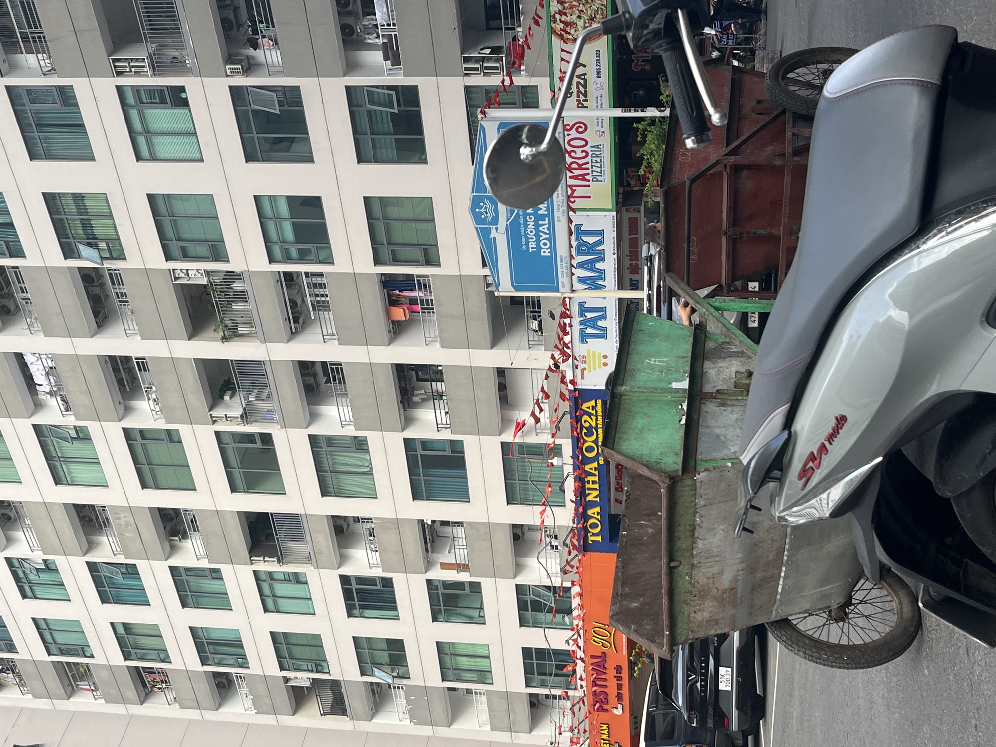 The image shows a street scene in Bac Nha Trang Ward, Vietnam, dominated by a large apartment building with multiple floors and balconies. In the foreground, several large metal trash receptacles are visible. A motorcycle is parked in the immediate foreground, partially obscuring the view of a dark-colored car parked to the left of the trash receptacles. Multiple businesses are advertised on banners and signs attached to the building at street level, including "FESTIVAL 90!", "TÒA NHÀ OC2A", "TAT MART", "MARCO'S PIZZERIA", and "ROYAL M...". Red and white pennant banners are strung across the street in front of the businesses. The sky is not visible due to the height of the building.