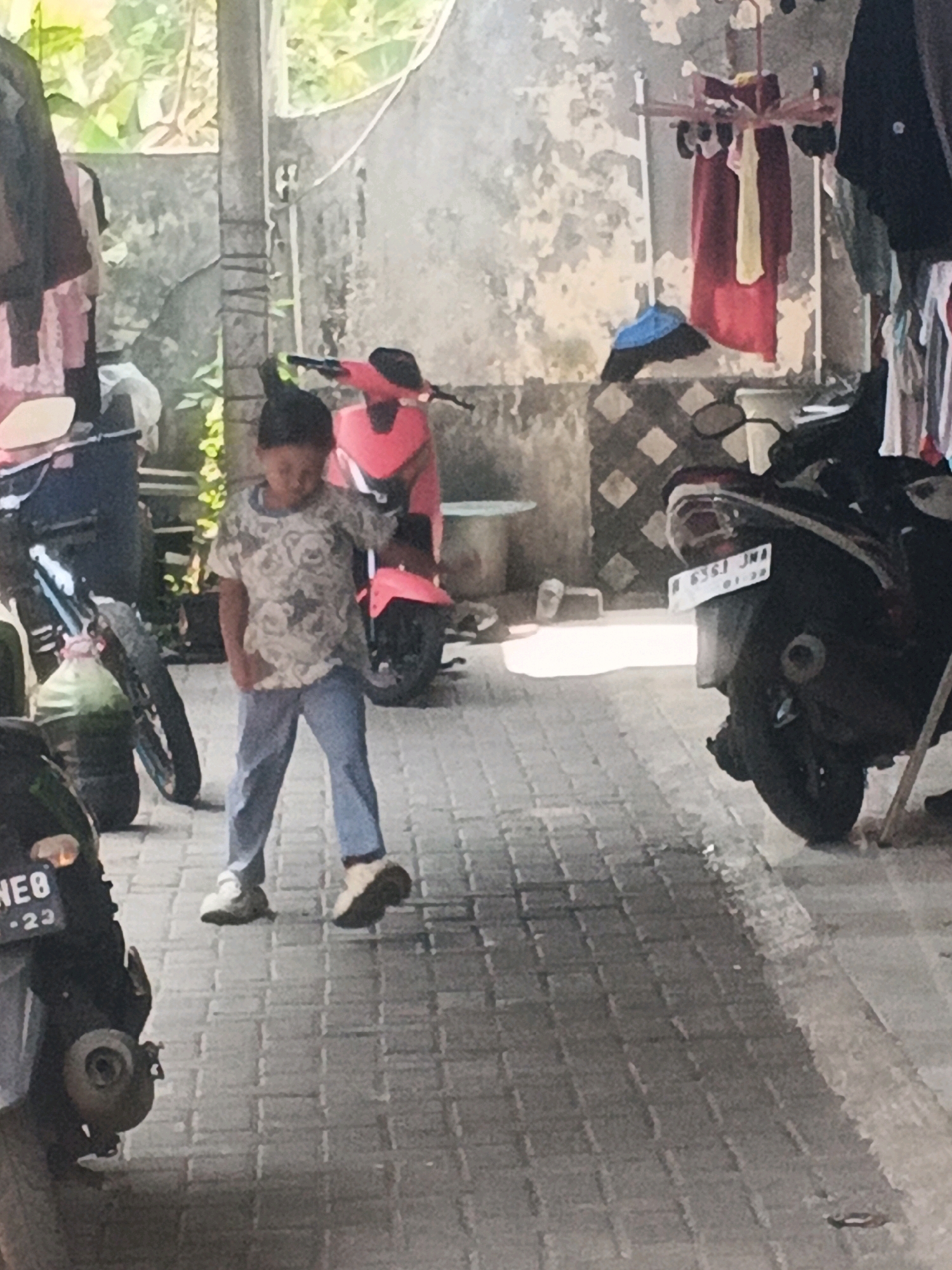 The image depicts a narrow outdoor pathway paved with brick-like stones. On the left, a portion of a black motorcycle is visible, with a license plate showing "NE0 -23." A young person, appearing to be a child, stands in the center of the pathway, facing towards the right and looking down. They are wearing a light-colored patterned t-shirt and blue pants, with white sneakers on their feet. To the right of the child, a pink scooter is parked. In the background, there is a textured wall, some greenery, and laundry hanging on a rack. Further to the right, another dark-colored motorcycle is parked, also with a visible license plate that reads "A 6361 JMA." The scene suggests an urban or semi-urban environment, potentially a residential area or a workshop, possibly located in Kadu, Indonesia, given the context.