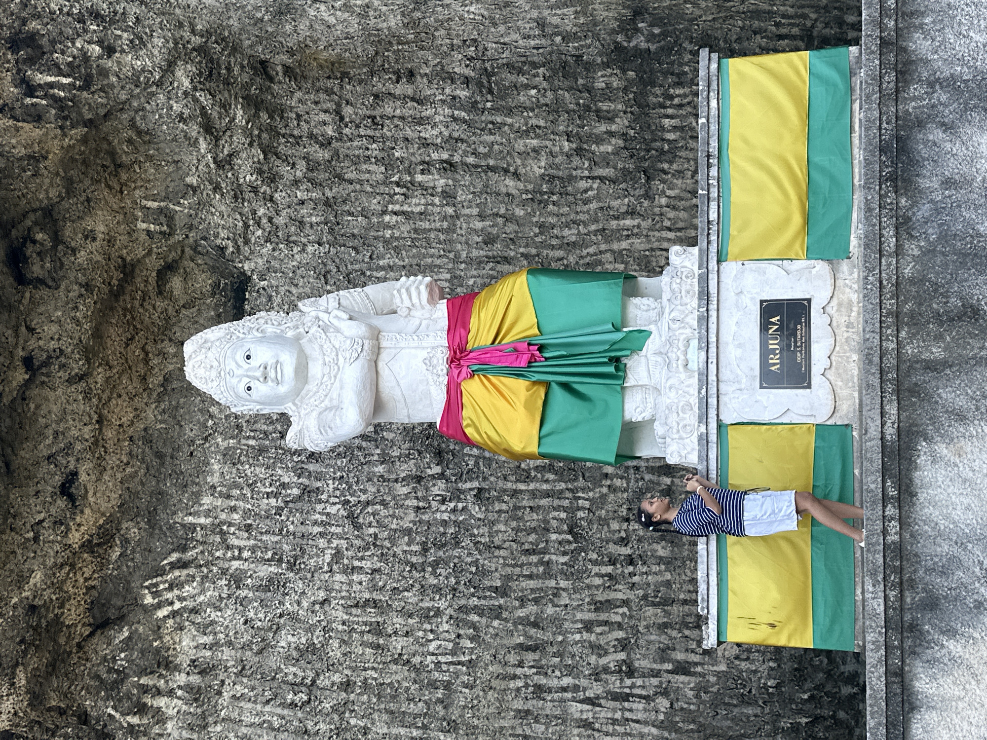 This media file shows a young girl lying on a yellow and green flag. She is wearing a striped blue and white shirt and white shorts. Behind her, a large white statue of a person is depicted, adorned with a pink, yellow, and green sash. The statue appears to be carved from stone and is set against a rough, grey rock face. The flag and the base of the statue are placed on a light-colored, textured surface, possibly a platform or walkway. In front of the statue, a plaque with the name "ARJUNA" is visible. The scene suggests a cultural or historical site. The lighting indicates it is daytime. The setting is outdoors, with the rough texture of the rock face suggesting a natural or man-made cave-like environment. The girl's posture and expression do not convey a strong emotion, appearing relaxed. The location is identified as Kuta Selatan, Indonesia.
