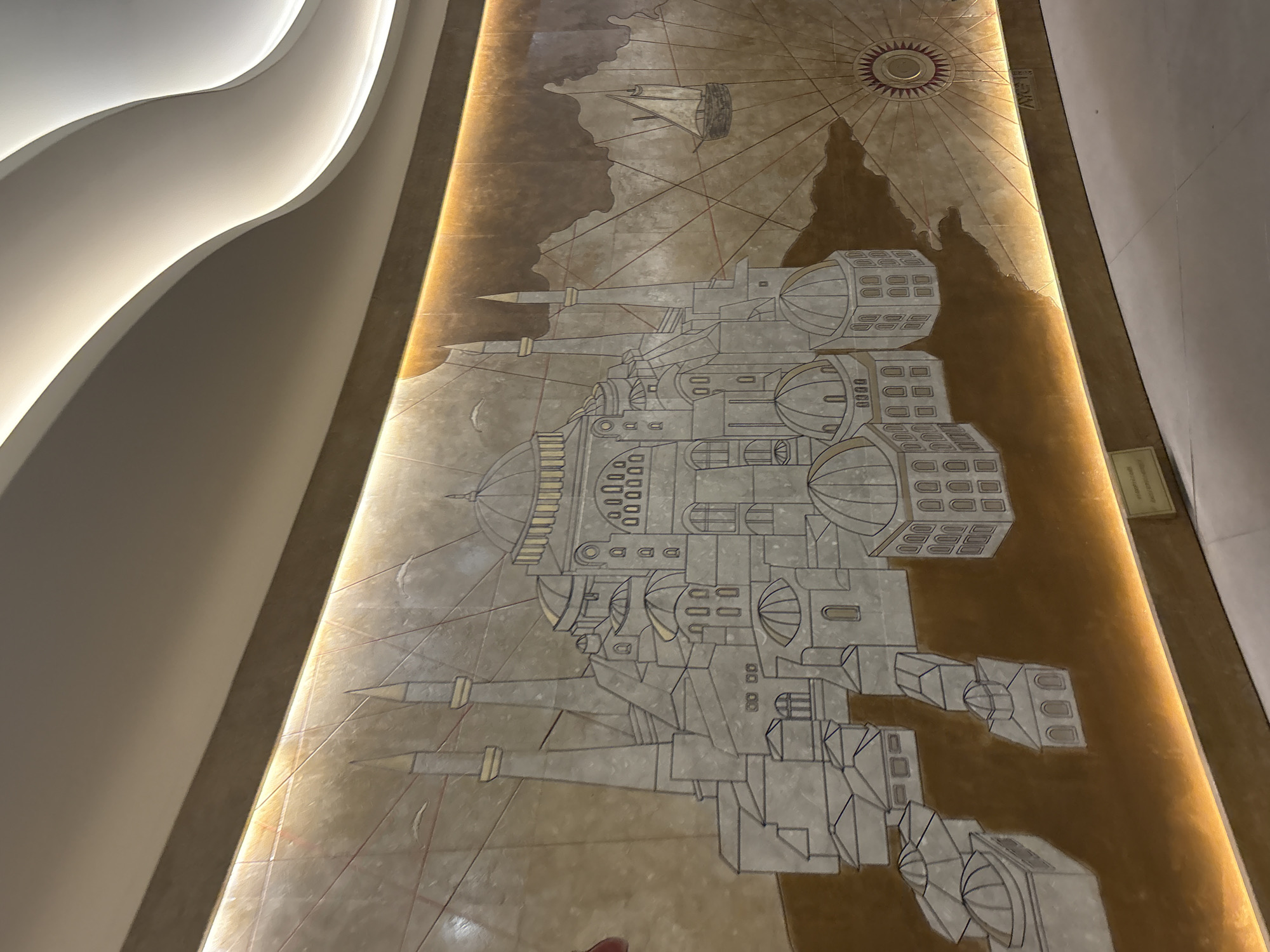 This image captures a section of an indoor wall featuring a large, ornate mural crafted from various light-colored and warm-toned inlaid materials, possibly stone or tile. The central subject of the mural is a detailed, stylized depiction of Hagia Sophia, rendered in shades of light gray and white with gold accents on its domes and minaret tops. The architectural representation is set against a map-like background in multiple shades of brown and gold, illustrating a landmass and featuring a single-masted sailing ship in the upper right quadrant. A series of radiating lines, originating from a central circular emblem resembling a compass rose in the lower right, overlays the background. The mural is framed by warm LED strip lighting running horizontally along both its top and bottom edges, illuminating the artwork and casting a glow on the adjacent surfaces. Above the mural, the ceiling exhibits a contemporary design with flowing, curvilinear white panels that incorporate concealed lighting, creating soft, indirect illumination. The floor visible at the bottom of the image is a plain, light-colored surface. A small, rectangular informational plaque is mounted on the wall below the mural, on the left side, though its text is not legible. No people are visible in the scene. This specific mural is located within the Mecidiyeköy station of the Istanbul Metro M7 line, situated in the Şişli district of Turkey.
