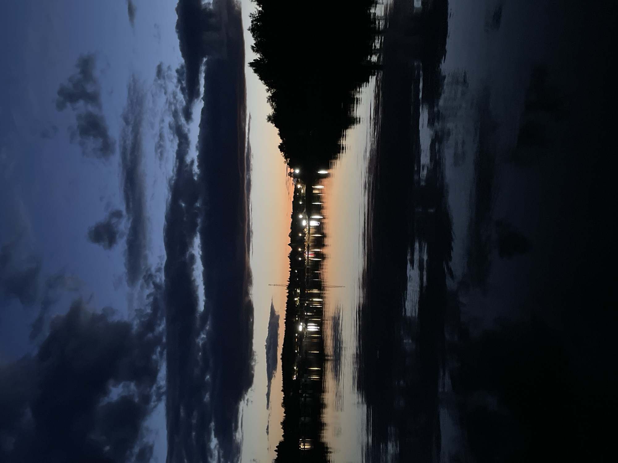 The image depicts a twilight scene over a body of water, likely a river or lake, in Berozovo, Russia. The sky is a deep blue transitioning to hues of orange and yellow near the horizon, with dark, dramatic clouds overhead. Silhouetted trees line the distant shore, punctuated by a scattering of lights from what appears to be a settlement or town. These lights are reflected in the calm water, creating shimmering patterns. The foreground is dominated by the dark water, reflecting the sky and the distant lights. No people or specific actions are visible, creating a still and atmospheric landscape.