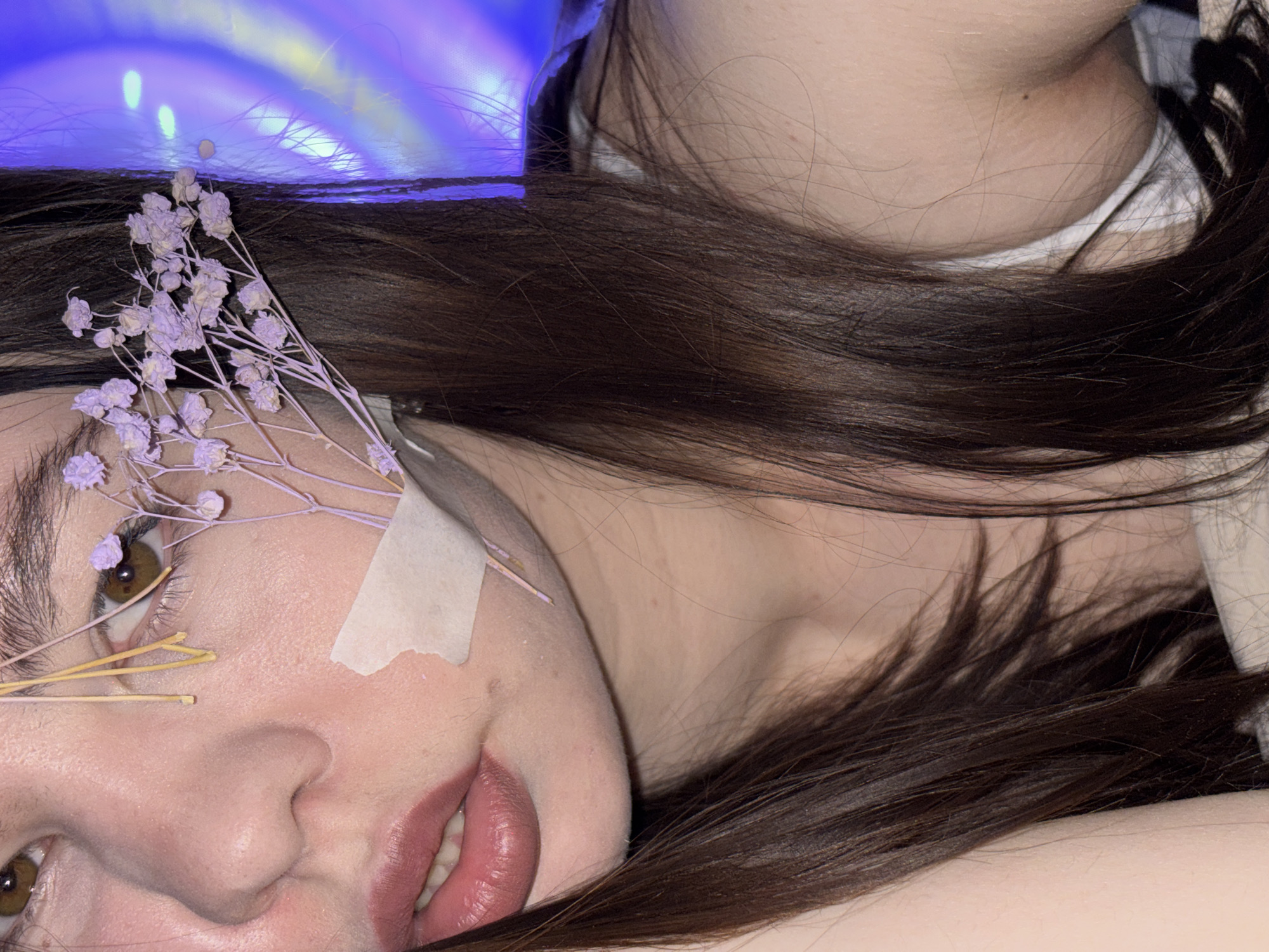 The image is a close-up, artistic portrait of a person (likely female) lying down in an indoor setting in Perm, Russia, under soft, artificial lighting. The primary subject's face is partially visible, angled upwards from the lower-left, with long, dark brown hair cascading across the frame and partially obscuring the lower right.
A delicate sprig of light purple dried flowers, resembling baby's breath, rests on their forehead and hair. Several thin, dried yellow strands are positioned near their left eye. A small piece of light-colored tape is affixed to their left temple/cheek, adding a stylistic or conceptual element. The subject's left eye, with a warm brown iris, is open and gazes towards the viewer. Their lips are slightly parted, revealing a hint of teeth and adorned with a muted pinkish-red lipstick.
In the upper-right portion, the smooth skin of another person's neck and shoulder is visible, with dark hair flowing over it, and a glimpse of a white garment or undergarment.
The background is a vibrant, abstract blur of blue and purple light, indicative of an artificial light source like neon or LED strips, creating an ethereal and atmospheric setting. The lighting suggests the photograph was taken in the evening or at night. The overall mood conveyed is one of tranquility, introspection, or artistic contemplation. No text is visible in the image.