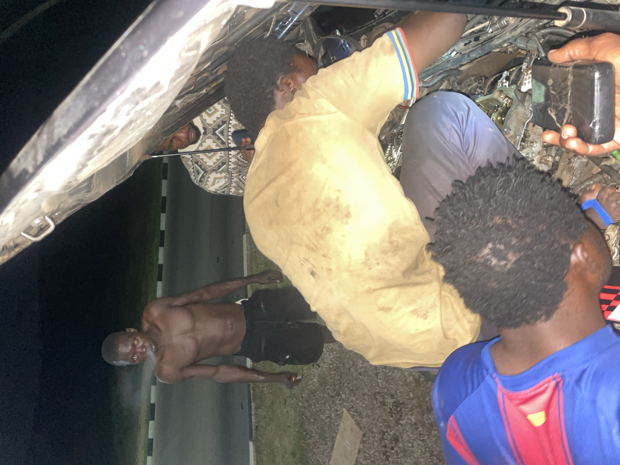 In a nighttime scene at Mopol 44 Barracks Kuje, Nigeria, three men appear to be working on the engine of a vehicle. One man, shirtless and wearing black shorts, is leaning against a road barrier. Another man, wearing a dirty yellow t-shirt with colorful stripes on the sleeves and blue pants, is actively engaged with the engine. A third man, partially visible and wearing a blue t-shirt with red and yellow accents, is also near the engine. He is holding a dark, rectangular object, possibly a phone or a tool. The vehicle's hood is open, revealing the complex engine components. The ground appears to be gravelly. The lighting suggests it is dark outside, with artificial light sources illuminating the scene. The men's expressions are not clearly discernible due to the lighting and their positions.