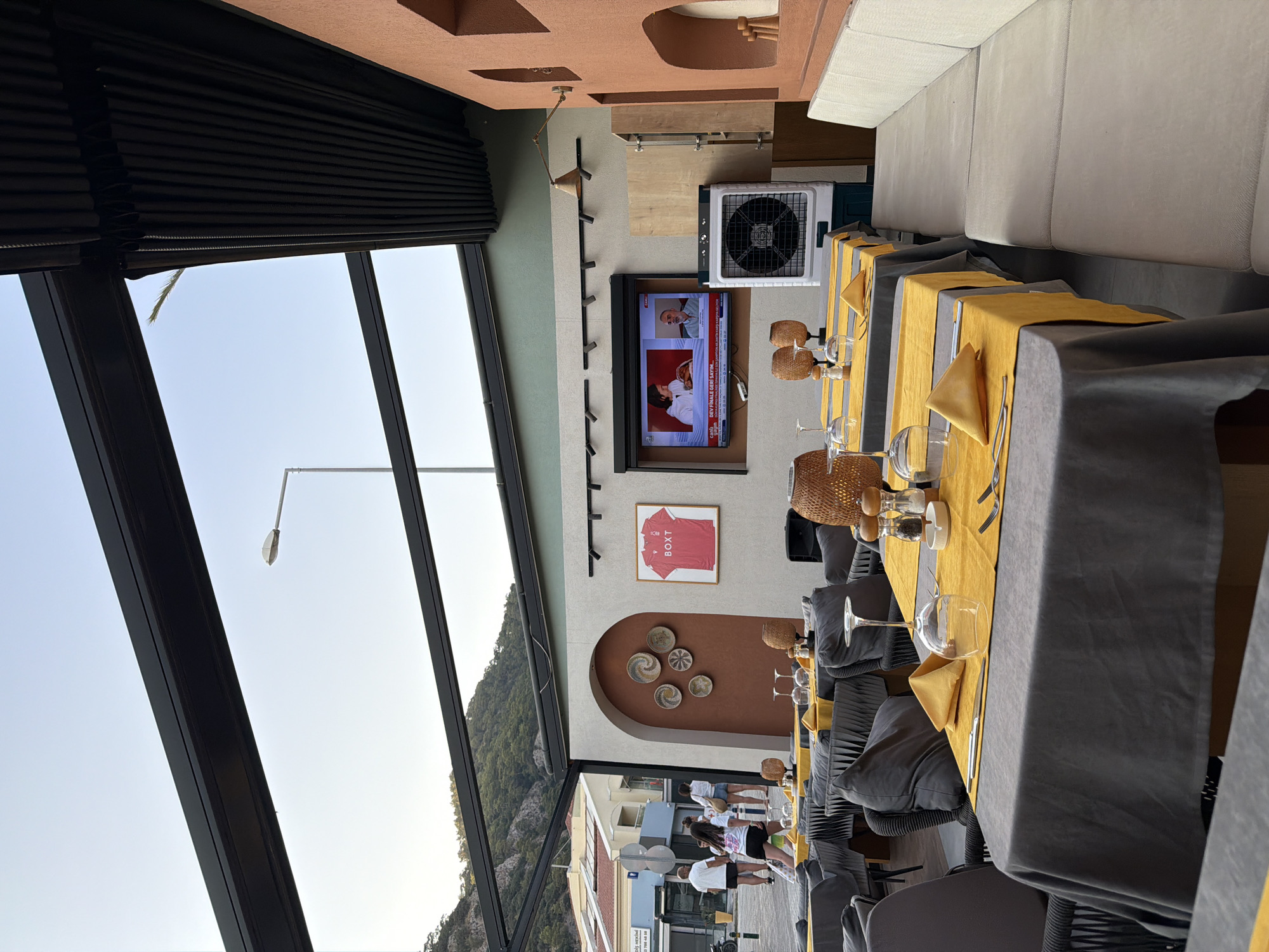 The image shows an outdoor seating area of a restaurant in Fethiye, Turkey. The seating area is covered by a retractable roof with black louvres. Tables are set with grey tablecloths, yellow placemats, and wine glasses. In the background, a television displays a news program. To the right, a large industrial fan is positioned near a wall with several decorative niches. A long, upholstered bench is visible on the far right. The environment suggests a warm climate, with the sky appearing clear and bright. The restaurant appears to be located on a street, as indicated by the presence of people walking in the background and buildings typical of a town or city.
