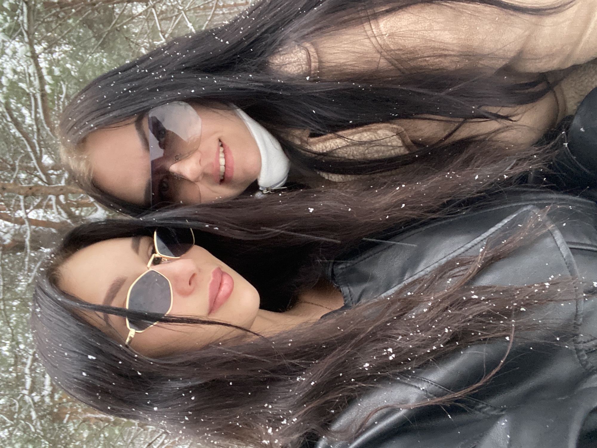 Two women with long, dark hair are positioned close together, facing the camera. They both wear sunglasses. The upper woman is smiling and wearing a tan jacket and a white turtleneck. The lower woman is wearing a black leather jacket. Snowflakes are visible in their hair and on their clothing, suggesting a winter setting. The background appears to be a wooded area with snow-covered branches. The photograph was taken in the city of Chaikovsky, Russia.