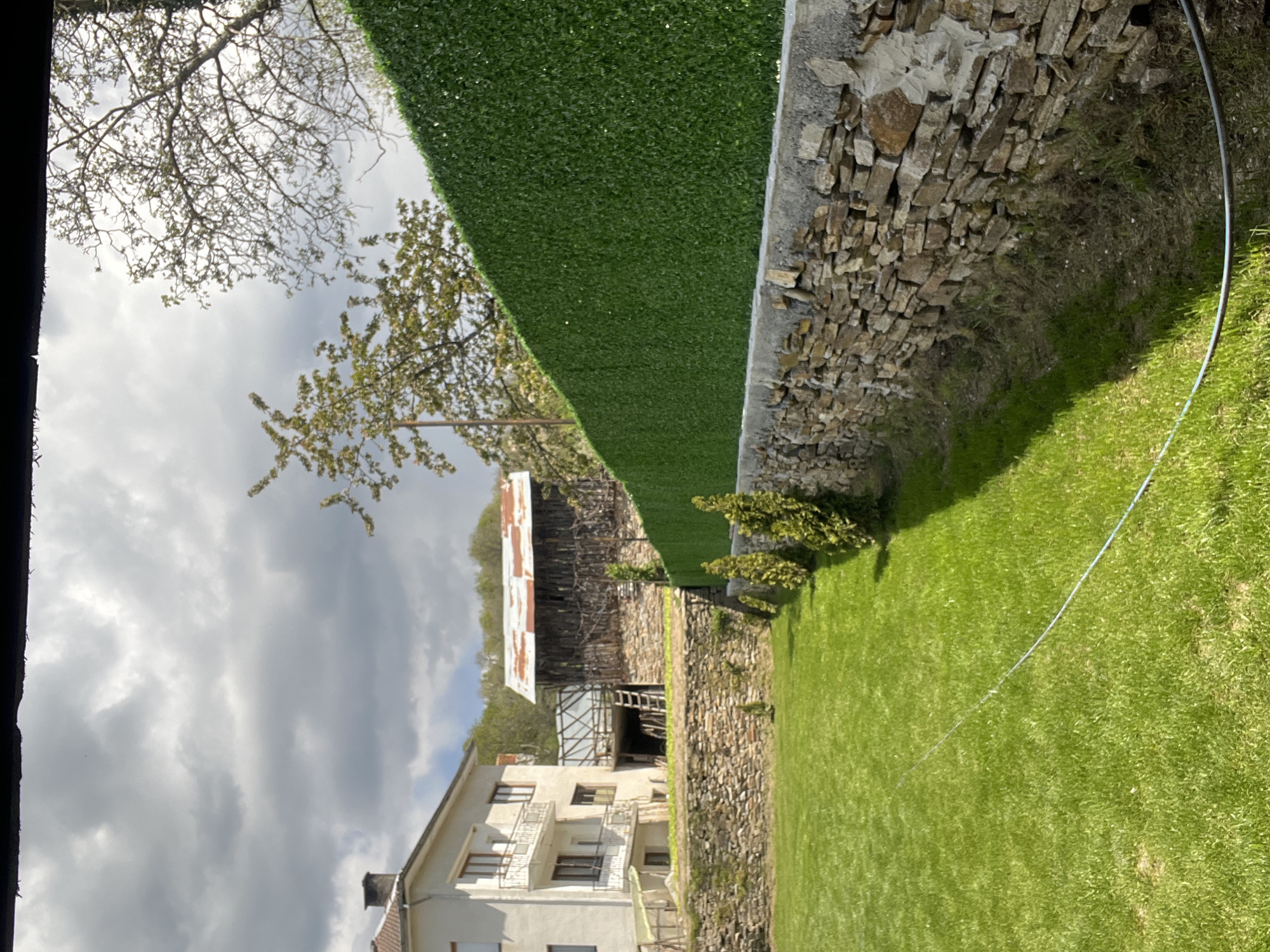 This image captures a quiet, sunlit rural or suburban landscape in Kribul, Bulgaria, viewed under an overcast yet bright sky. The scene is static, with no visible human activity.
In the foreground, a lush, well-maintained green lawn stretches across the lower and right portions of the frame. A thin, light-colored cable or hose lies on the grass, extending from the right edge towards the center. Separating the lawn from a lower area is a sturdy stone wall, built with natural, irregularly shaped rocks, running horizontally across the mid-ground. Perched atop this stone wall is a neat, uniform artificial green hedge or fence, providing a verdant backdrop.
To the left of the stone wall and below the level of the lawn, a multi-story house with light-colored facades and multiple balconies is visible. Further back and to the right of this house, behind the artificial hedge, stands a rustic, older building, possibly a barn or outbuilding. This structure features exposed wooden beams and a large, partially rusted corrugated metal roof. A few small, leafy bushes or trees are clustered near the stone wall on the lower ground level.
The background reveals a mix of trees; some are full of green leaves, while others show more bare branches against the sky. The sky above is predominantly cloudy, with varying shades of gray and white, suggesting an overcast day with some bright light filtering through. A dark, vertical frame element partially obscures the far left edge of the image. No text is visible anywhere in the scene.
