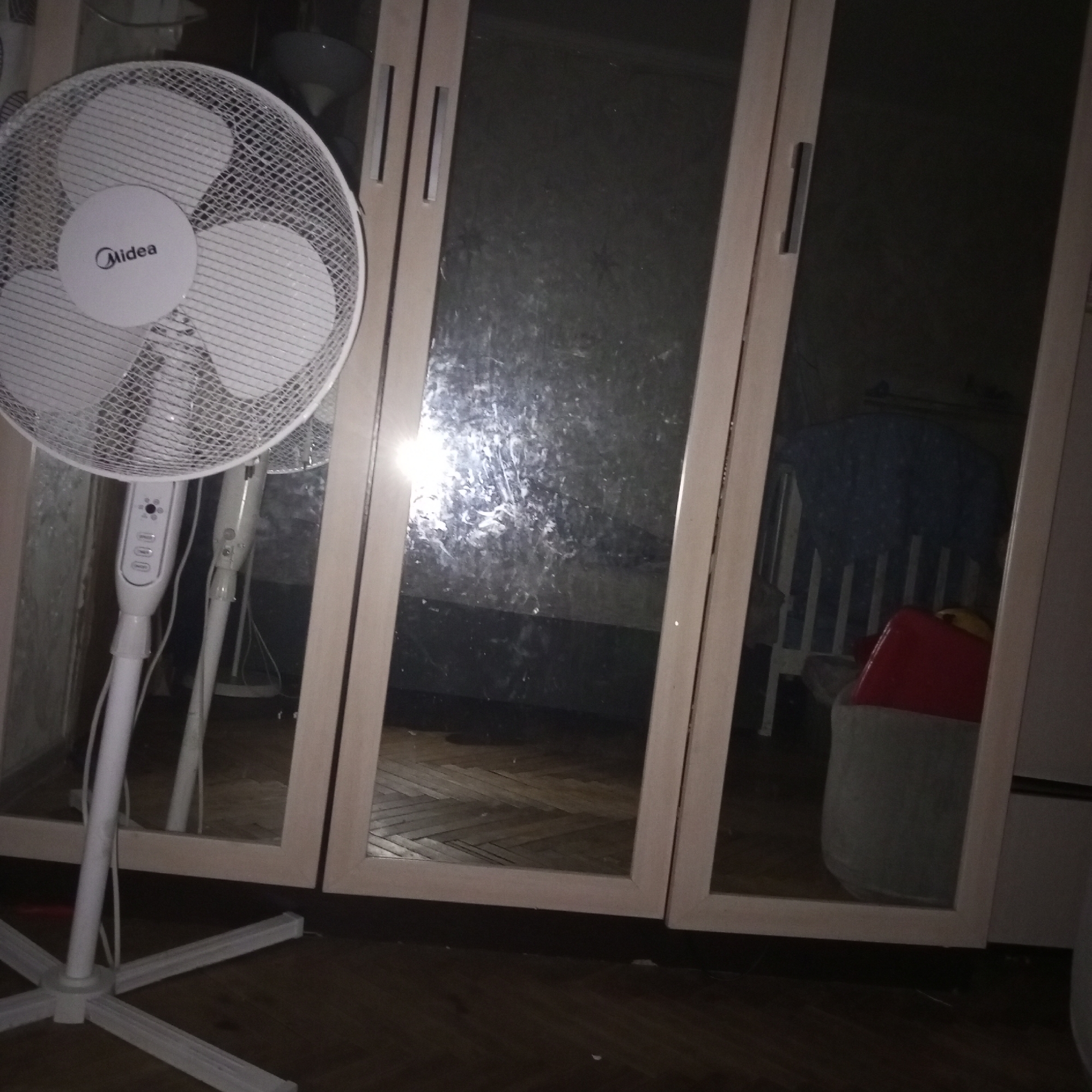 The image shows a standing fan in the foreground, with its base and pole clearly visible. Behind the fan, there are three mirrored closet doors. The middle and right closet doors reflect a room with a blue patterned object, possibly a child's bed or crib, and a red object that could be a toy or container. The left closet door reflects the fan and the surrounding environment. The flooring appears to be wood. The lighting is dim, suggesting an indoor setting, possibly a bedroom or storage area. The scene does not provide any specific indicators of its location in Moscow, Russia.