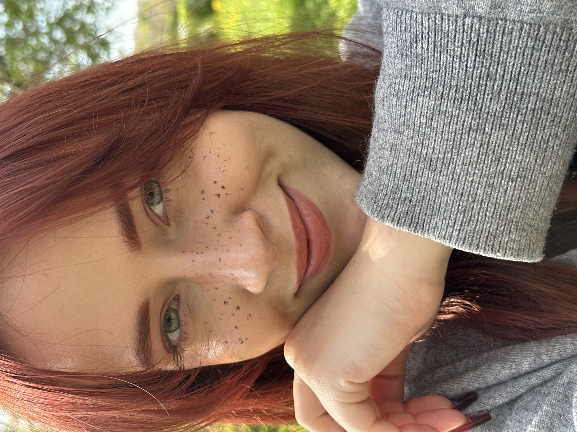 A close-up, tilted portrait shows a woman with auburn hair and light skin. She has freckles on her nose and cheeks, and her eyes are green. She is wearing a gray sweater, and her hand, adorned with long, dark nails, rests on her face. The background is blurred, showing indistinct greenery. The location is Unknown, Unknown.