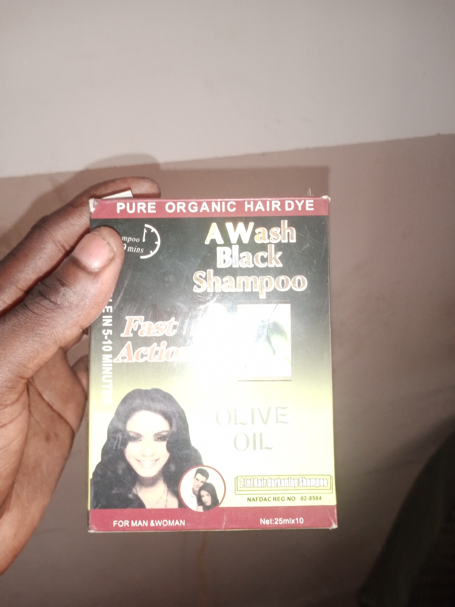 A hand is holding a box of "AWash Black Shampoo" hair dye. The box is red and gold, with text indicating "PURE ORGANIC HAIR DYE" and "Fast Action". It features an image of a woman's face and a smaller inset image of a couple. The text "OLIVE OIL" is prominent on the box, along with "2 in 1 Hair darkening Shampoo" and "NAFDAC REG. NO: 02-8584". The box also states "Net: 25mlx10" and "FOR MAN & WOMAN". The background is a plain, light-colored wall. This image was taken in Jalingo, Nigeria.