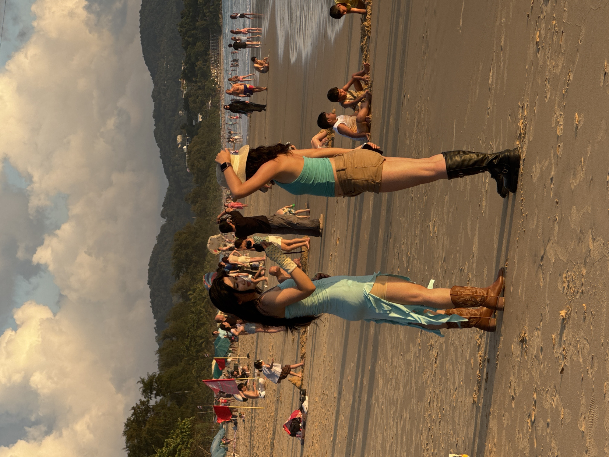 Two women stand on the sand at Karon Beach, Thailand. The woman on the left wears a light blue dress with cutouts and brown cowboy boots. The woman on the right wears a teal tank top, brown shorts, and black boots. She holds a straw hat in her right hand, which is raised to her head. The beach is crowded with people, some wading in the ocean and others sitting on the sand. Palm trees and hills line the background. The sky is filled with large, fluffy clouds.