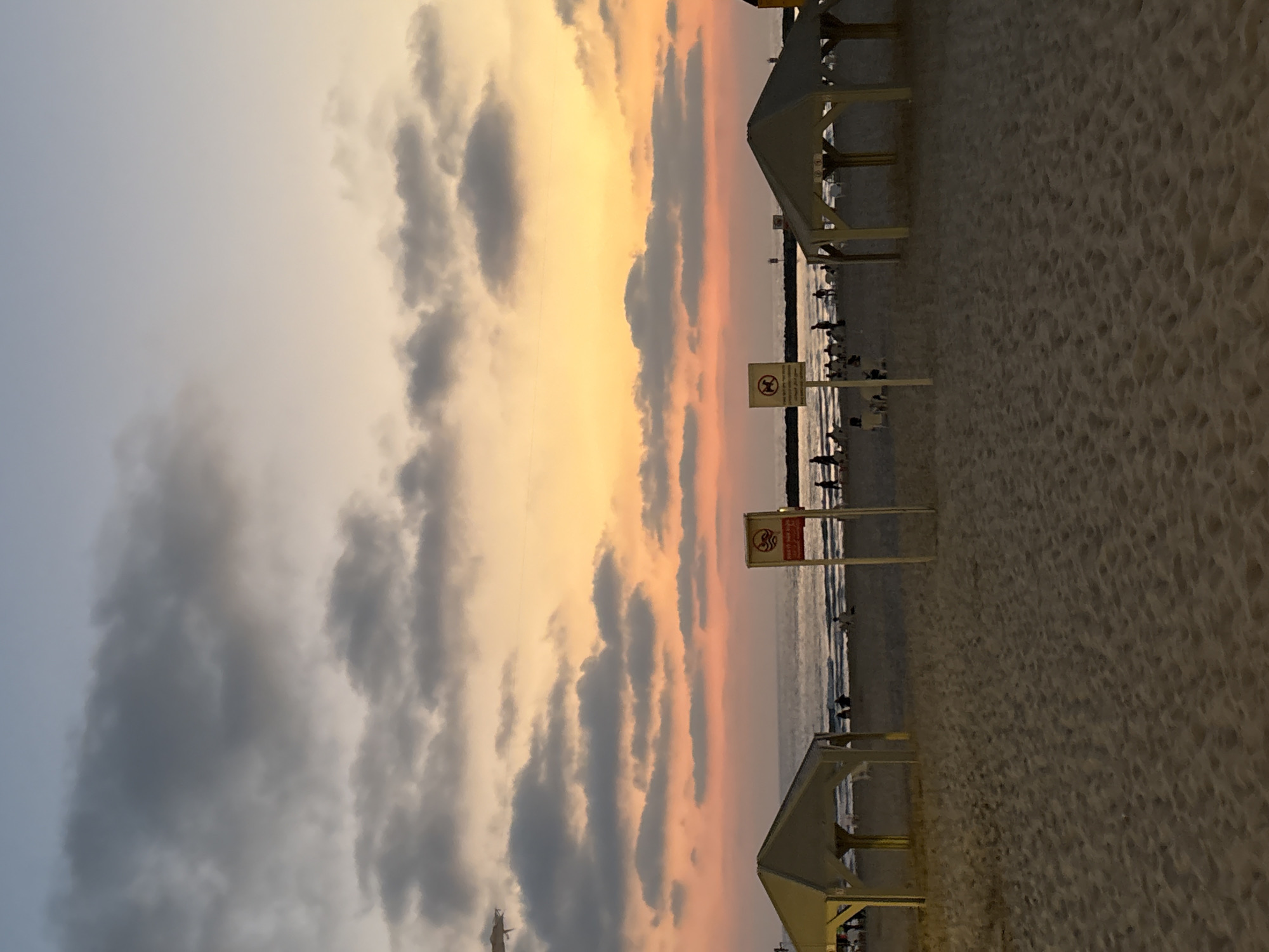 This is a photograph of a beach in Tel Aviv, Israel, taken at sunset. The sky is filled with warm hues of orange, pink, and yellow, with scattered clouds. In the foreground, there is a sandy beach with two small, light-colored cabanas. People are visible in the distance, walking along the shoreline and sitting on the sand. The ocean waves are gently lapping at the shore. Two signs are posted on poles on the beach, one with a red border and a symbol of a person swimming, and another with a red border and a symbol of a dog crossed out. There appears to be an airplane in the upper left corner of the sky. The overall scene is tranquil and depicts a typical evening at a popular beach.