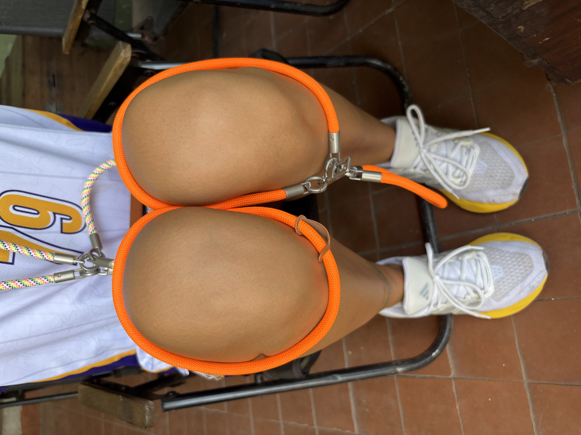 The image shows a close-up of a person's knees and legs, viewed from above. The person is wearing a white sports jersey with yellow and purple accents, and the number "79" is visible in yellow. Their knees are bent, and an orange rope is looped around them, secured by metal clasps. The person is also wearing white athletic shoes with yellow soles. The background shows a tiled floor and parts of outdoor chairs, suggesting an outdoor or semi-outdoor setting. The lighting is bright, indicating daylight. The scene appears to be a personal moment or activity. The location is not identifiable from the image itself.