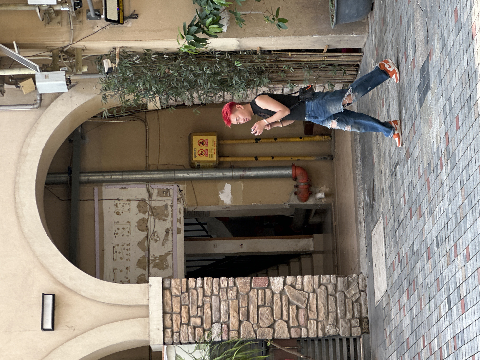 A person with red hair is looking at a phone in an alley in Chongqing High-tech Zone, China. The person is wearing a black tank top and ripped blue jeans with orange sneakers. The alley has an arched entrance, a brick wall, and a tiled floor. There are pipes running along the wall. The sky is visible at the top right of the image.
