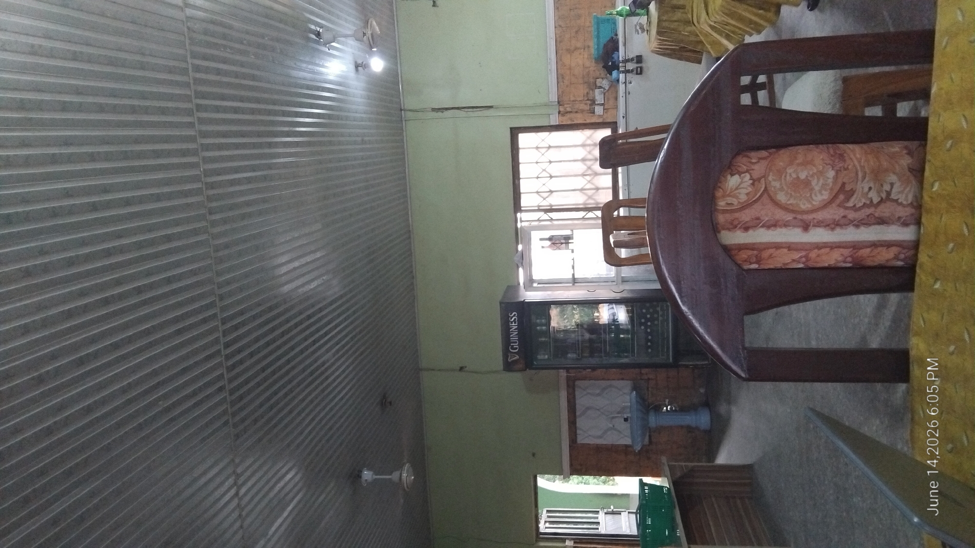 The image depicts the interior of a building, possibly a bar or restaurant, located in Howayi, Nigeria. The ceiling is made of corrugated metal sheeting. A white ceiling fan is visible on the left side of the image, and another on the right. The walls are painted a light green.
In the center of the room, a black commercial refrigerator with the "GUINNESS" logo is present. To the right of the refrigerator, there is a window with a metal security grate. Below the window, a long, white refrigerator or counter is visible, with a blue plastic crate on top.
In the foreground, a dark wooden chair with an ornate patterned cushion is prominent. Another wooden chair is visible behind it. A portion of a table with a yellow patterned tablecloth is in the lower right corner.
To the left of the Guinness refrigerator, there is a doorway leading outside, with a green basket on the floor just inside. A sink on a blue pedestal is situated against a brick-patterned wall.
The floor appears to be tiled. The overall impression is of a functional, somewhat dated establishment. A timestamp "June 14, 2026 6:05 PM" is overlaid on the bottom left of the image. There are no people clearly visible in the image.