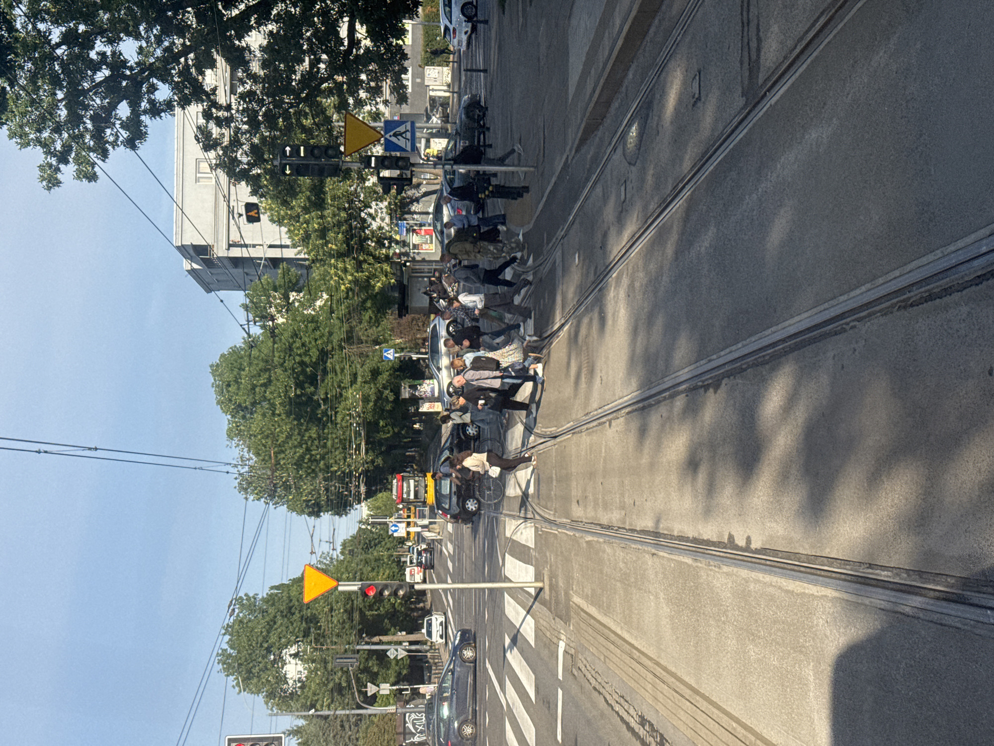 The image captures a street scene in Warsaw, Poland, likely at an intersection with tram tracks. Several pedestrians are crossing a busy street at a marked crosswalk. Cars are also present, stopped at the traffic light or waiting for pedestrians to cross. In the background, buildings and trees line the street. The sky is clear and blue, indicating a sunny day. The tram tracks are prominently visible in the foreground, suggesting this is a common route for public transportation. The overall atmosphere is that of a typical urban environment during daylight hours.