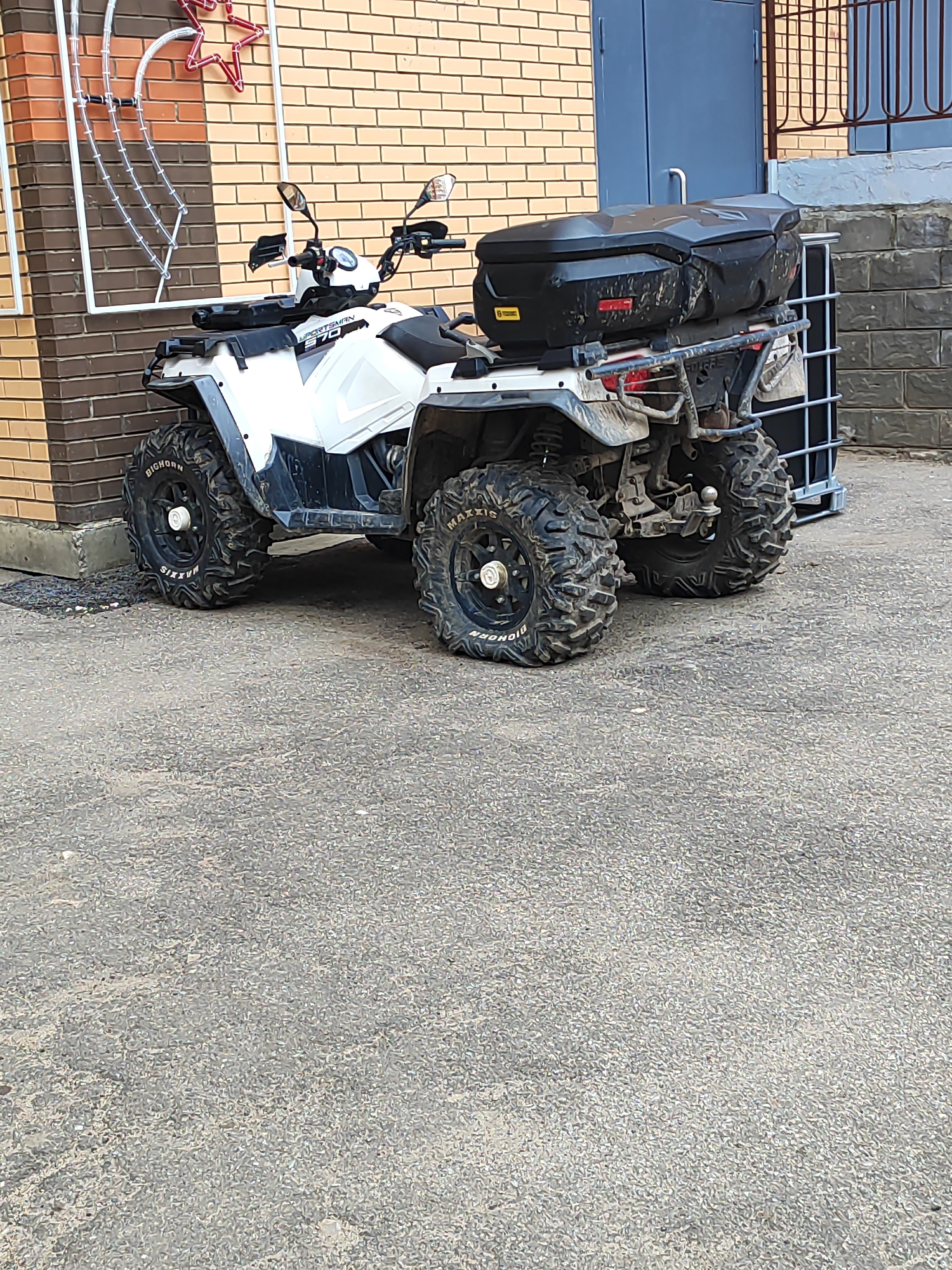 The image depicts an all-terrain vehicle (ATV) parked on a paved surface. The ATV is white and black, with "Sportsman 570" markings visible. It features large, knobby tires labeled "Maxxis Bighorn." A black storage box is mounted on the rear of the ATV. The ATV is situated next to a brick building with a decorative star-shaped light fixture. A dark gray door and a wire fence are also visible in the background. A container made of metal bars is behind the ATV. The location is Shchyolkovo, Russia, based on the GPS data available in the image metadata.