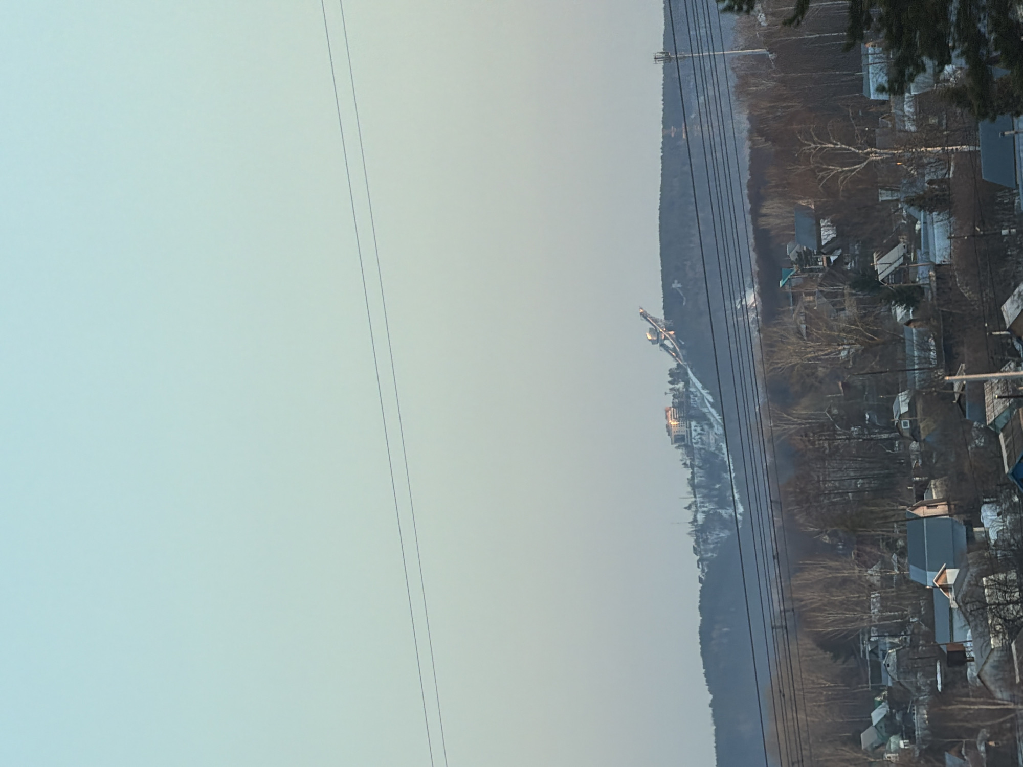 The image presents a panoramic view of a residential and natural landscape in Noviy, Russia, under a clear, pale blue sky.
In the foreground, a dense cluster of detached houses with varied roof colors (predominantly grey and blue-grey) are visible, interspersed with bare deciduous trees and patches of snow-covered ground. Multiple dark, horizontal power lines traverse the mid-ground, partially obscuring the view.
The mid-ground consists of a band of bare deciduous trees and some evergreens, also with snow visible on the ground.
In the background, a large, snow-dusted forested hill or mountain dominates the horizon. Two prominent structures are situated on this distant hill: on the left, a large ski jump facility with an inclined ramp and a tall support tower; on the right, a multi-story building featuring reflective surfaces on its upper levels that display a distinct, bright golden-orange sheen, suggesting direct sunlight. A tall, slender communication tower is also visible on the right side of the hill.
The scene is illuminated by natural daylight, consistent with a winter or early spring environment due to the bare trees and snow. No individuals, specific actions, or interactions are discernible within the frame.