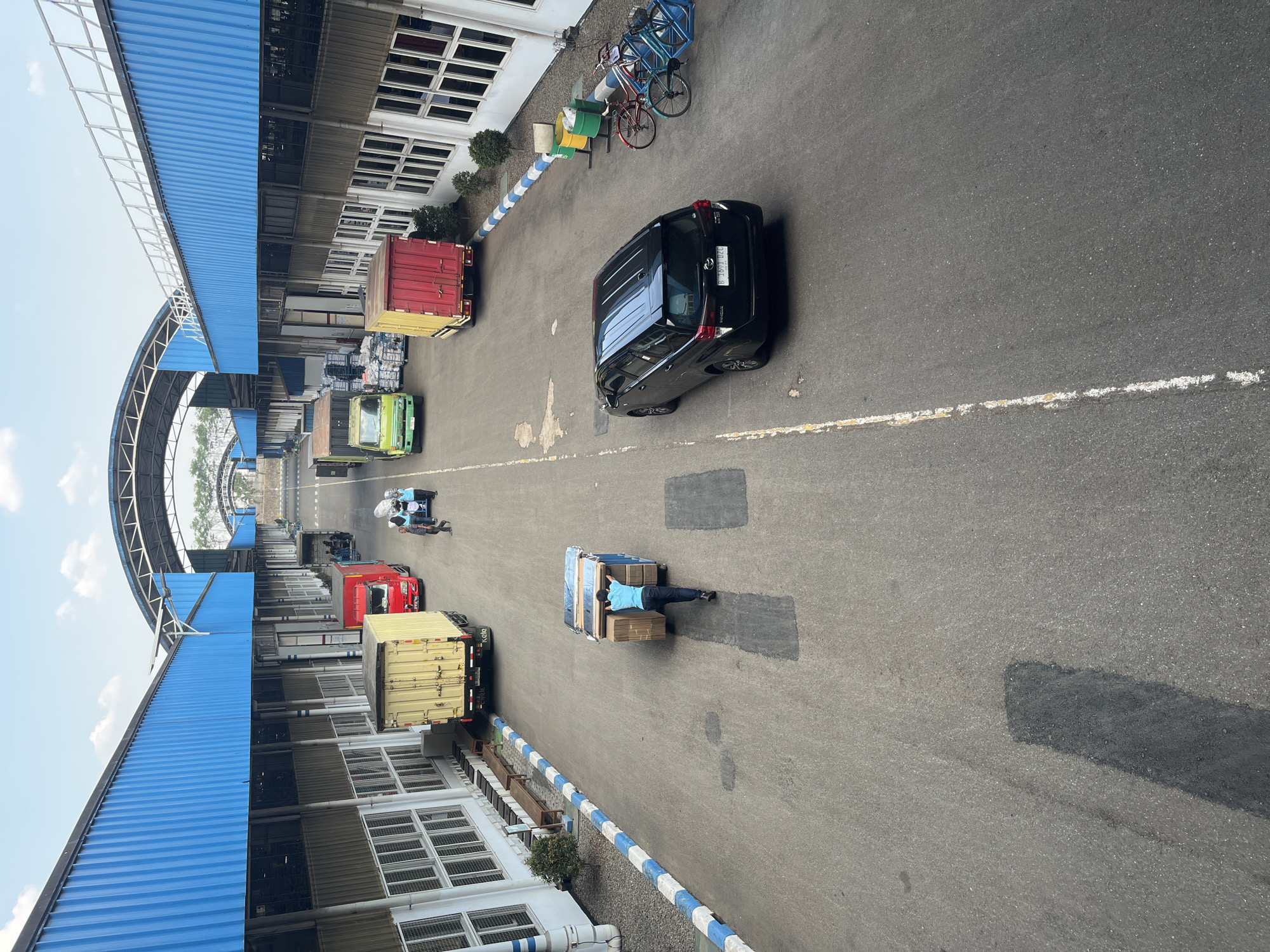 An overhead shot captures a bustling scene at a logistics facility located in Pelang, Indonesia. The image is dominated by a large, paved road flanked by industrial buildings with prominent blue roofs. Several trucks are parked or in motion on the road, including a yellow container truck, a red truck, and a green truck. In the foreground, a person in a blue shirt is pushing a cart laden with boxes. A black SUV is parked on the right side of the road. Two bicycles are propped up near some trash bins. The sky above is a light blue with scattered white clouds, suggesting daylight. The overall environment appears to be a functional and active shipping or distribution center.