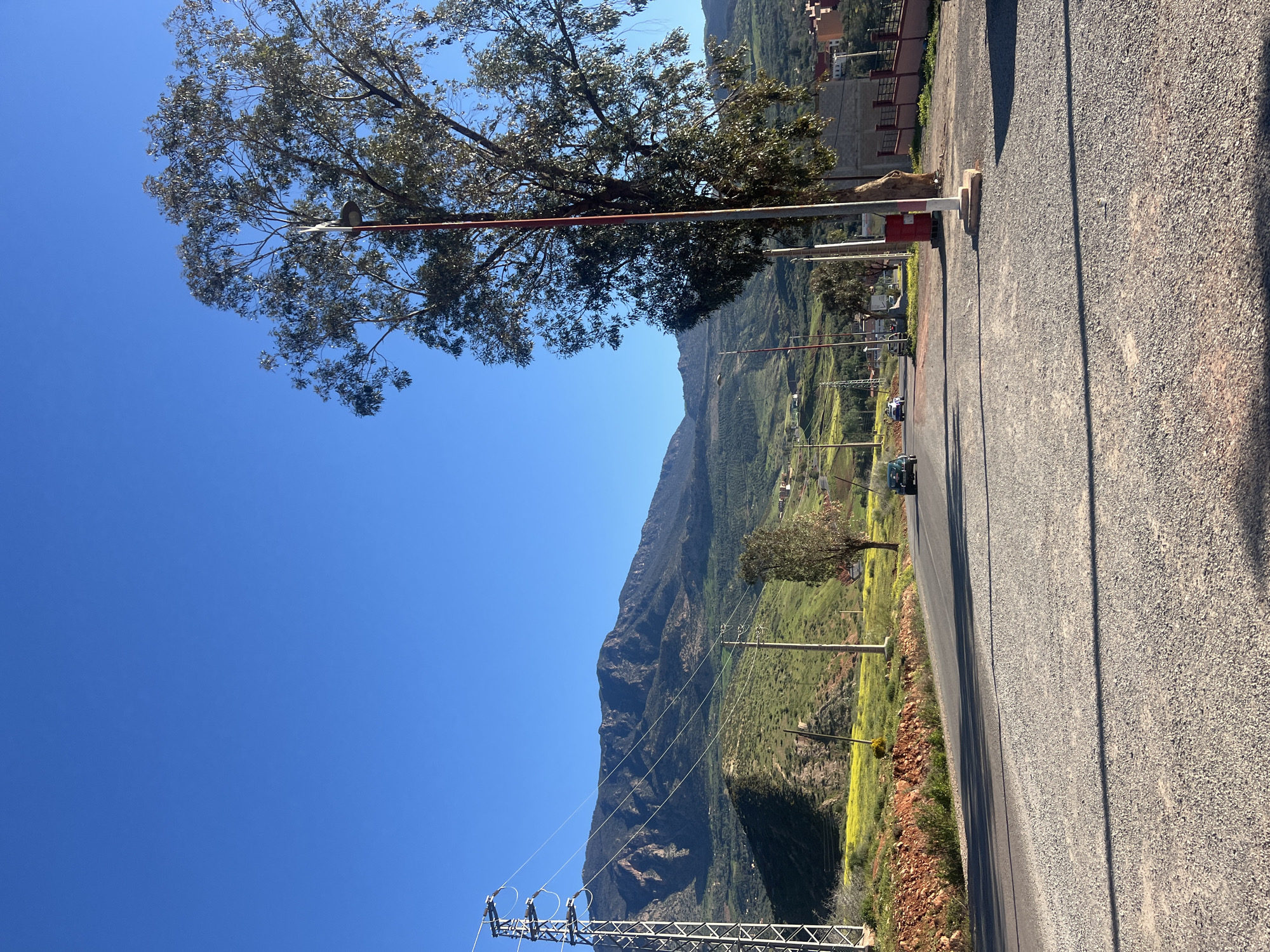 The image depicts a sunny, elevated view of Ouaouizert, Morocco, featuring a mountain range as a backdrop. A road winds through a green, mountainous landscape with scattered trees and power lines. A gate with a red and white barrier stands near a tree, suggesting an entrance or checkpoint. A vehicle can be seen driving along the road. The foreground shows a paved or gravel surface, likely a road shoulder. The sky is clear and blue. The photo is rotated counter-clockwise.