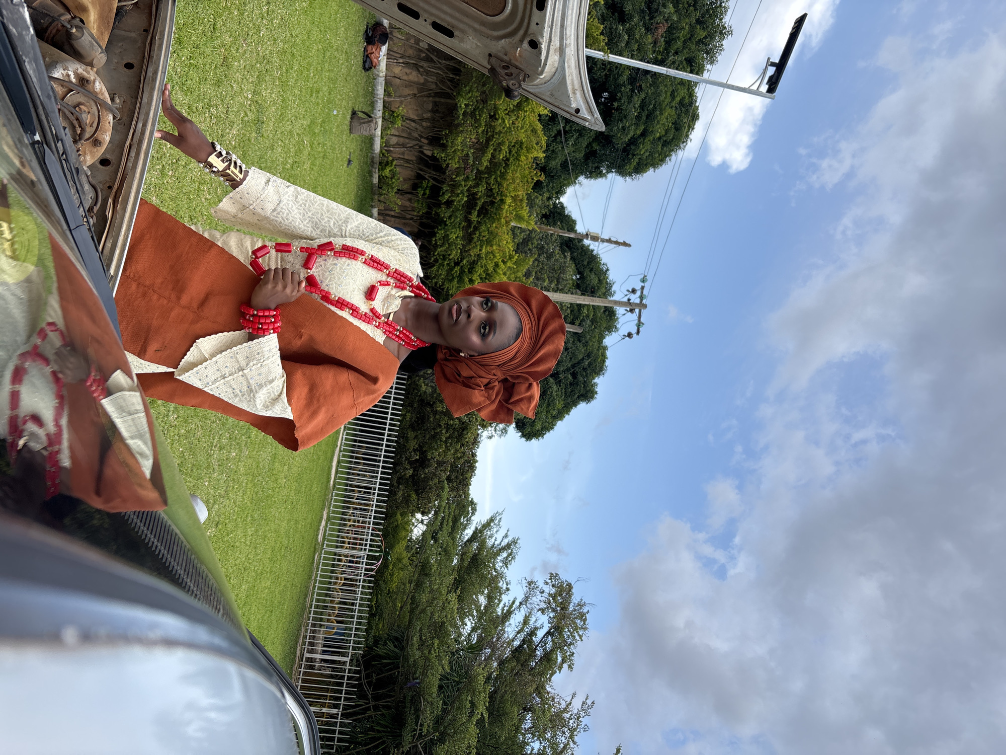 A full-body, low-angle outdoor shot captures a young Black woman posing next to an open car engine in what appears to be a residential area or a park in Jos, Nigeria. The woman is dressed in traditional attire, featuring a white lace top with ornate sleeves, an orange wrap, and a matching headscarf. She wears striking red beaded necklaces and bracelets, along with gold and pearl bangles. Her left hand rests on the edge of the open car hood, which reveals a visible engine block with various mechanical components. The background consists of a meticulously manicured green lawn, lush trees, a white metal fence, and a cloudy sky. The overall scene suggests a cultural or fashion photoshoot, possibly highlighting traditional Nigerian heritage against a contemporary urban backdrop.