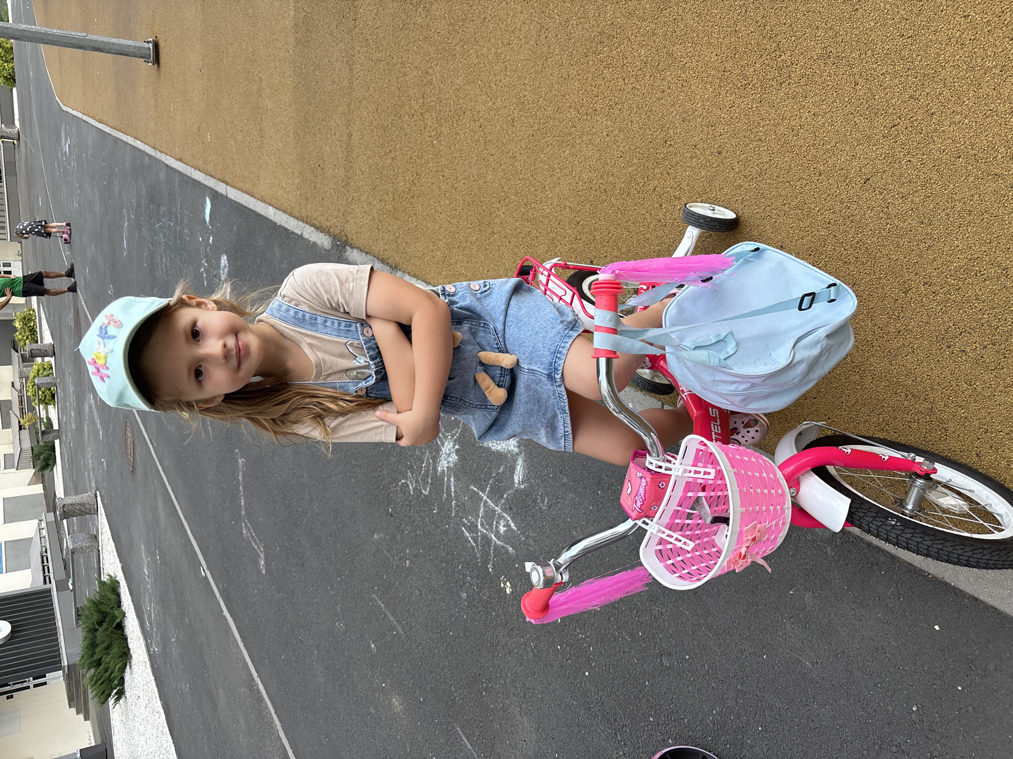 A young girl with long blonde hair and a light blue cap sits on a pink bicycle. She is wearing denim overalls over a light brown t-shirt and has her arms crossed. The bicycle has a white basket adorned with pink ribbons and a stuffed toy hangs from her overalls. A light blue bag is attached to the handlebars. The girl is on an asphalt surface with chalk drawings, adjacent to a yellowish-brown rubberized play area. In the background, two adults and another child are visible, with buildings and green bushes behind them. The location is the city of Orenburg, Russia.