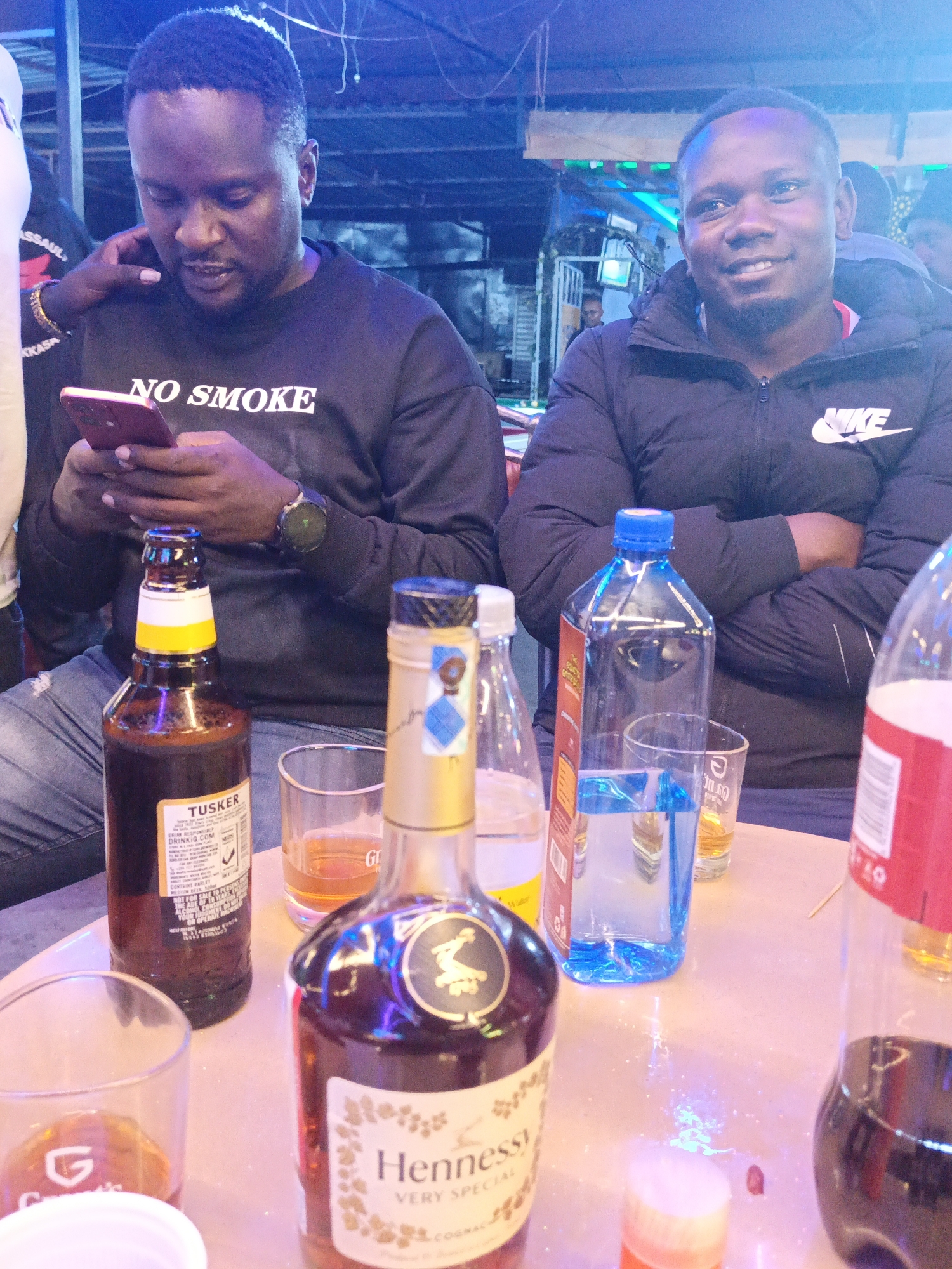 The image depicts two African men seated at a table covered with assorted alcoholic and non-alcoholic beverages in what appears to be a bar or club. The location is possibly in Nairobi, Kenya, given the presence of Tusker beer, a popular Kenyan brand. The man on the left is looking at his phone while wearing a black sweater emblazoned with the words "NO SMOKE." The man on the right is wearing a black Nike jacket with his arms crossed and smiling. A Hennessy bottle is prominently displayed on the table, along with other bottles of water, beer, and soft drinks. There are also partially filled glasses and a small toothpick on the table. The lighting is dim, with blue and purple hues creating a nightclub atmosphere. Other people are visible in the background, further suggesting a crowded public space.