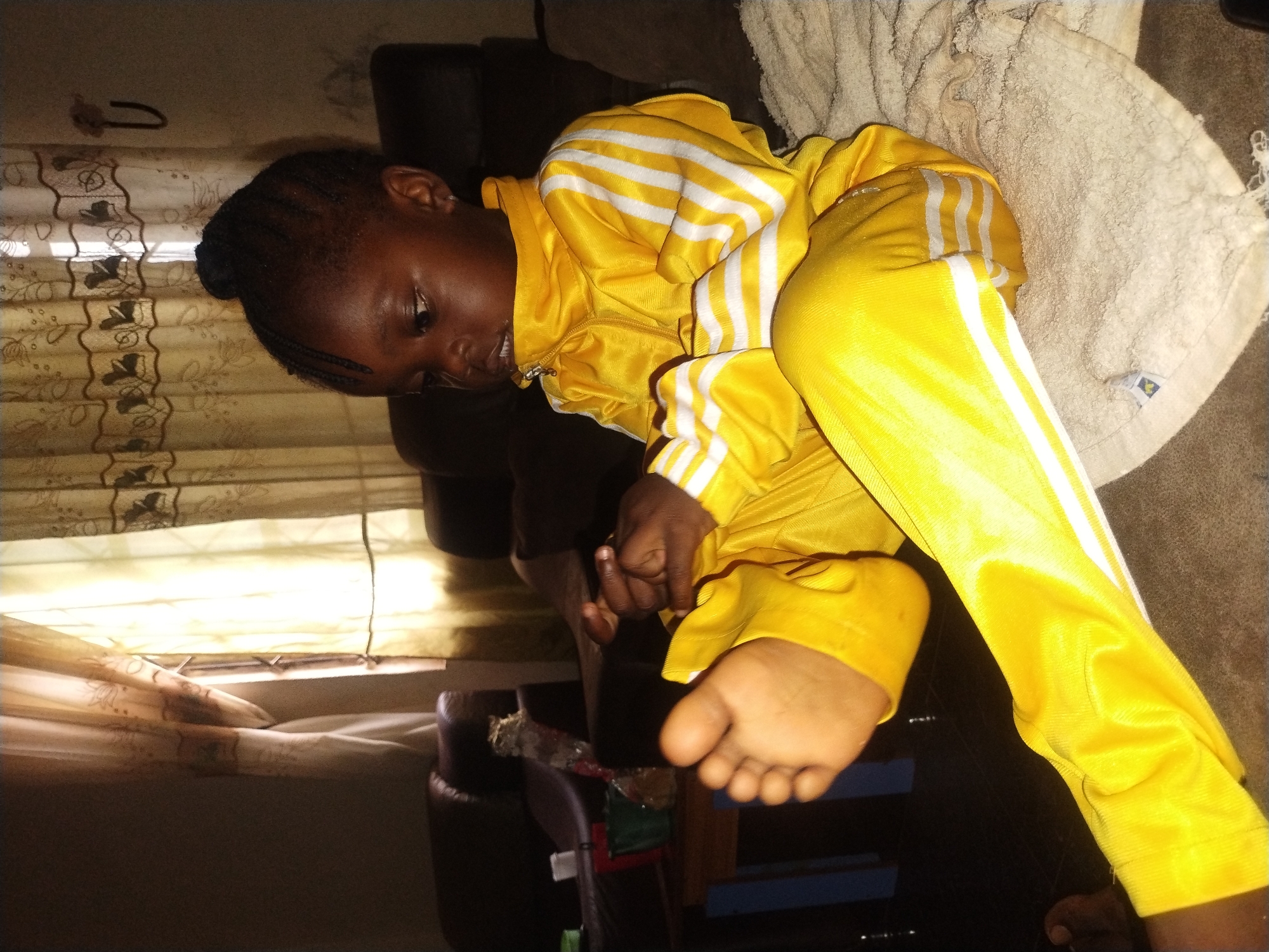 A young child with dark skin lies on a brown, upholstered chair. The child is dressed in a yellow tracksuit with white stripes on the sleeves and legs. The child's feet are bare and visible. The child is partially covered by a light-colored blanket. The background includes a window with patterned curtains. This photo was taken in Jos, Nigeria.