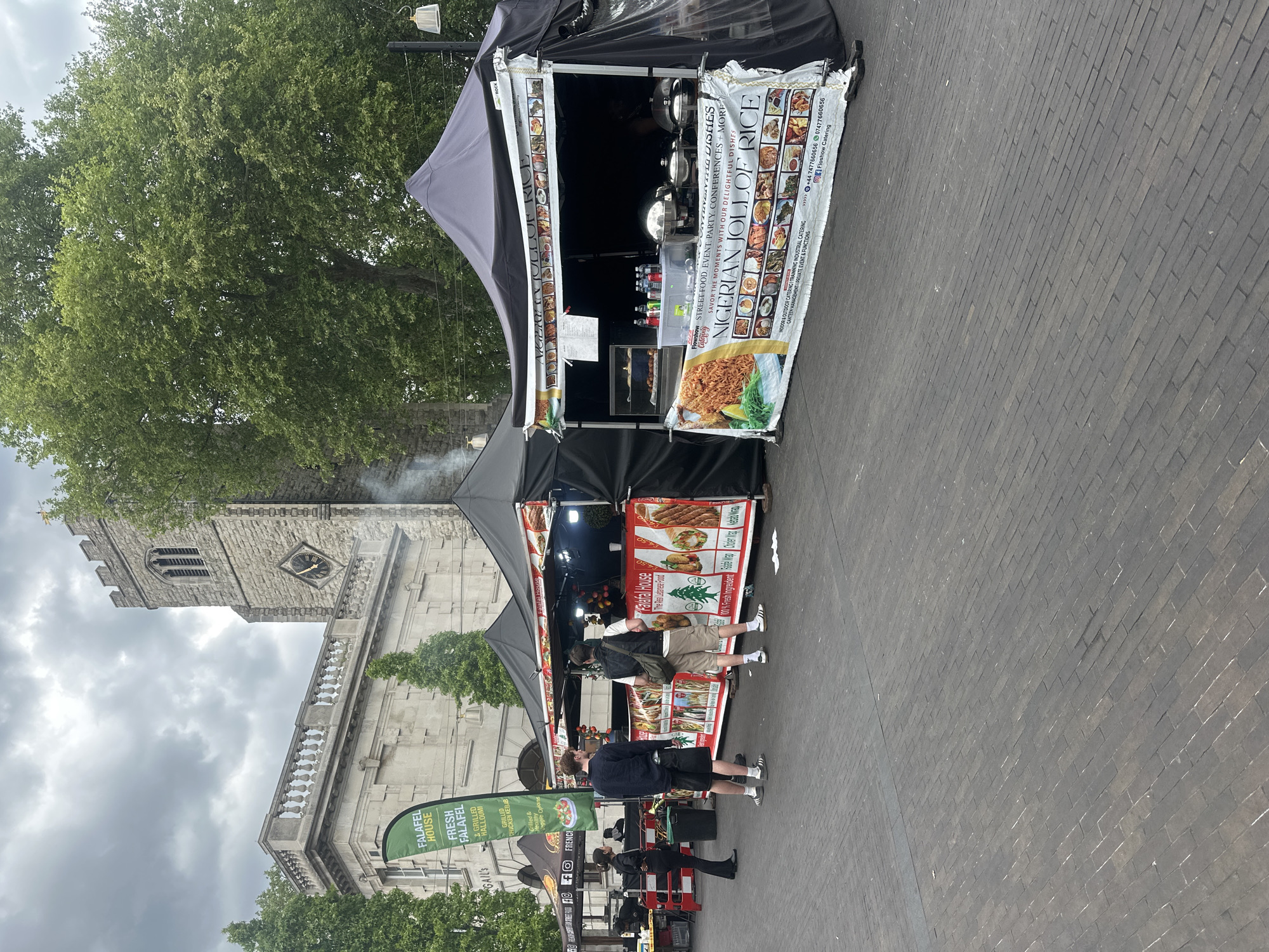 Here is a forensic description of the image:
The image captures an outdoor scene, likely a market or fair, with two food stalls being the focal point. The stall on the left is branded "Falafel House" and shows images of Lebanese food like falafel and kebabs. Two people, dressed in casual clothes, appear to be customers at this stall. The stall on the right is "Nigerian Jollof Rice" with a banner showing various dishes, as well as contact details.
Behind the stalls stands a gray stone building, its style indicating it may be a church or other historic structure. A banner that says "Gails" is partially visible. Above the building are trees with green foliage and a cloudy sky.
The ground is paved with gray blocks in a herringbone pattern. The scene appears to be set in London, United Kingdom, based on the style of the buildings and the market environment.