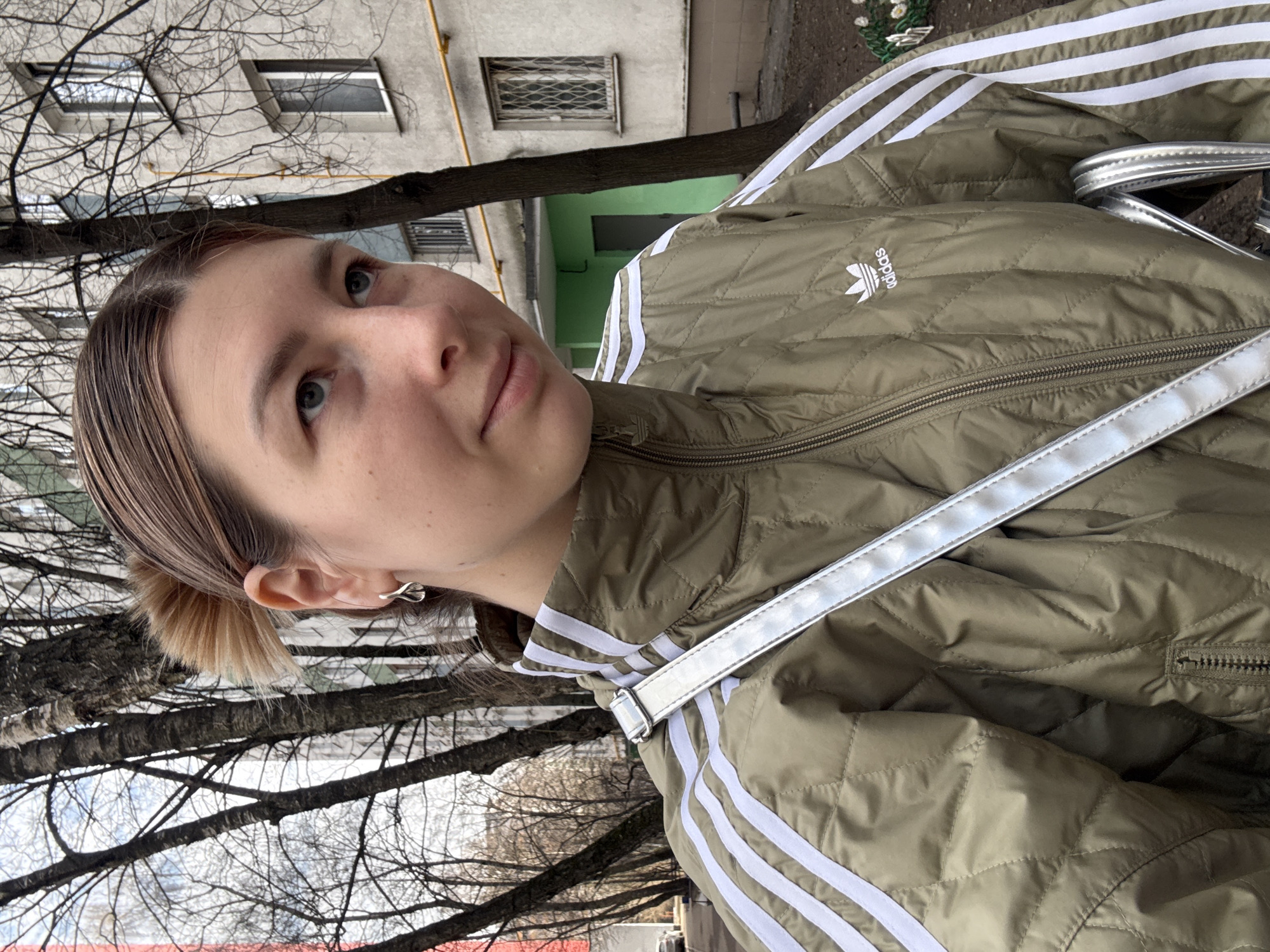 A woman stands outdoors in Moscow, Russia. She is wearing an olive green Adidas jacket with white stripes and a silver bag strap is visible. The building in the background is a multi-story apartment building with windows, some covered with metal grates, and a green door frame. Bare tree branches are visible in the foreground.