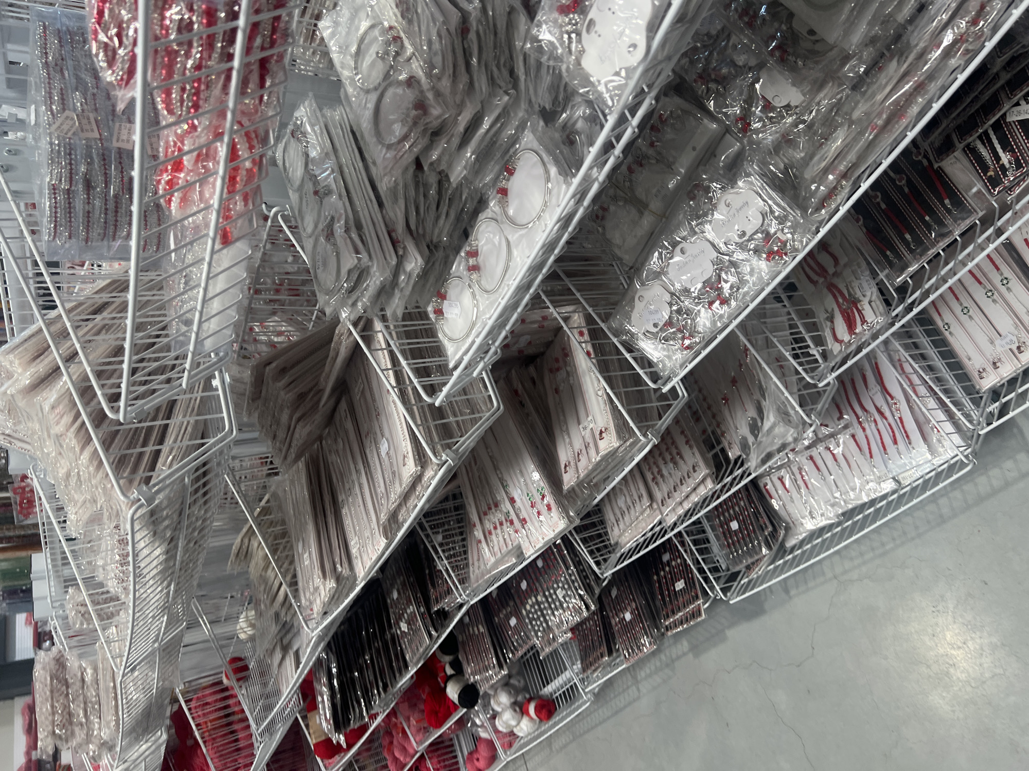 The image shows a commercial display of jewelry items in a retail setting. The jewelry is predominantly packaged in clear plastic and arranged on tiered white wire shelves. There is a wide variety of jewelry visible, including items with silver, red, and black components. Some packages contain multiple individual pieces. The shelves are densely packed with inventory, and the view is angled from above, looking down towards the floor. The floor is plain and light grey. The location appears to be a wholesale or retail store, possibly in Burgas, Bulgaria.