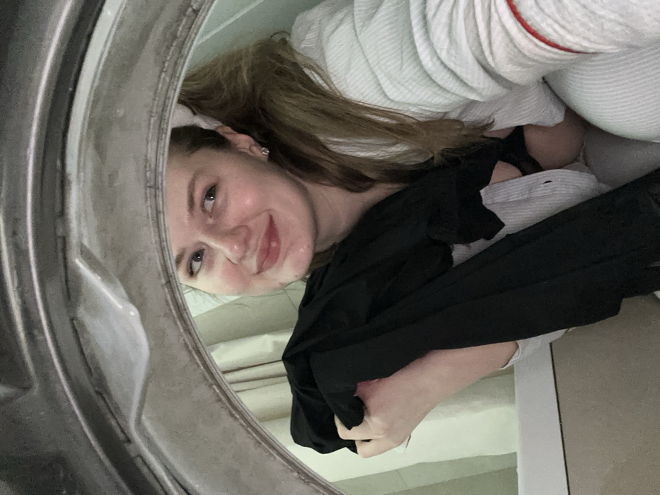 A young woman peers into the camera from inside a washing machine, holding black clothing in her hands. She has light skin, blonde hair, and a slight smile. The inside of the washing machine door is visible in the foreground. A striped shirt and white, quilted bathrobe are also present. The image is taken at an unusual angle, looking up into the washing machine from below.
