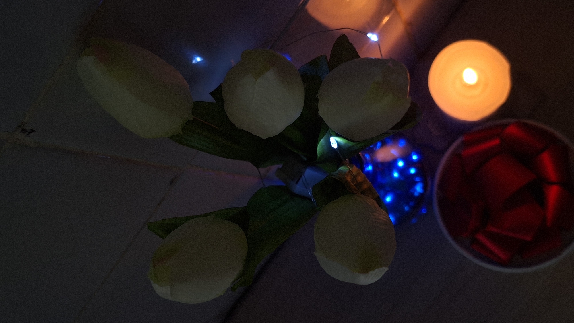 The image shows a still life composition with artificial tulips, a red gift box, and a lit candle, set against a dark background. The tulips, likely made of fabric or plastic, are a pale yellow color and are arranged in a clear vase. They are partially illuminated by fairy lights, which are predominantly blue, with some white and orange lights also visible. The gift box is round and red, appearing to contain a red ribbon. The candle is circular and glows with a warm orange light. The scene is lit by the candle and the fairy lights, creating a low-light, atmospheric setting. The location of Забалоцкі сельскі Савет, Belarus is not discernible from the visual information provided in the image.