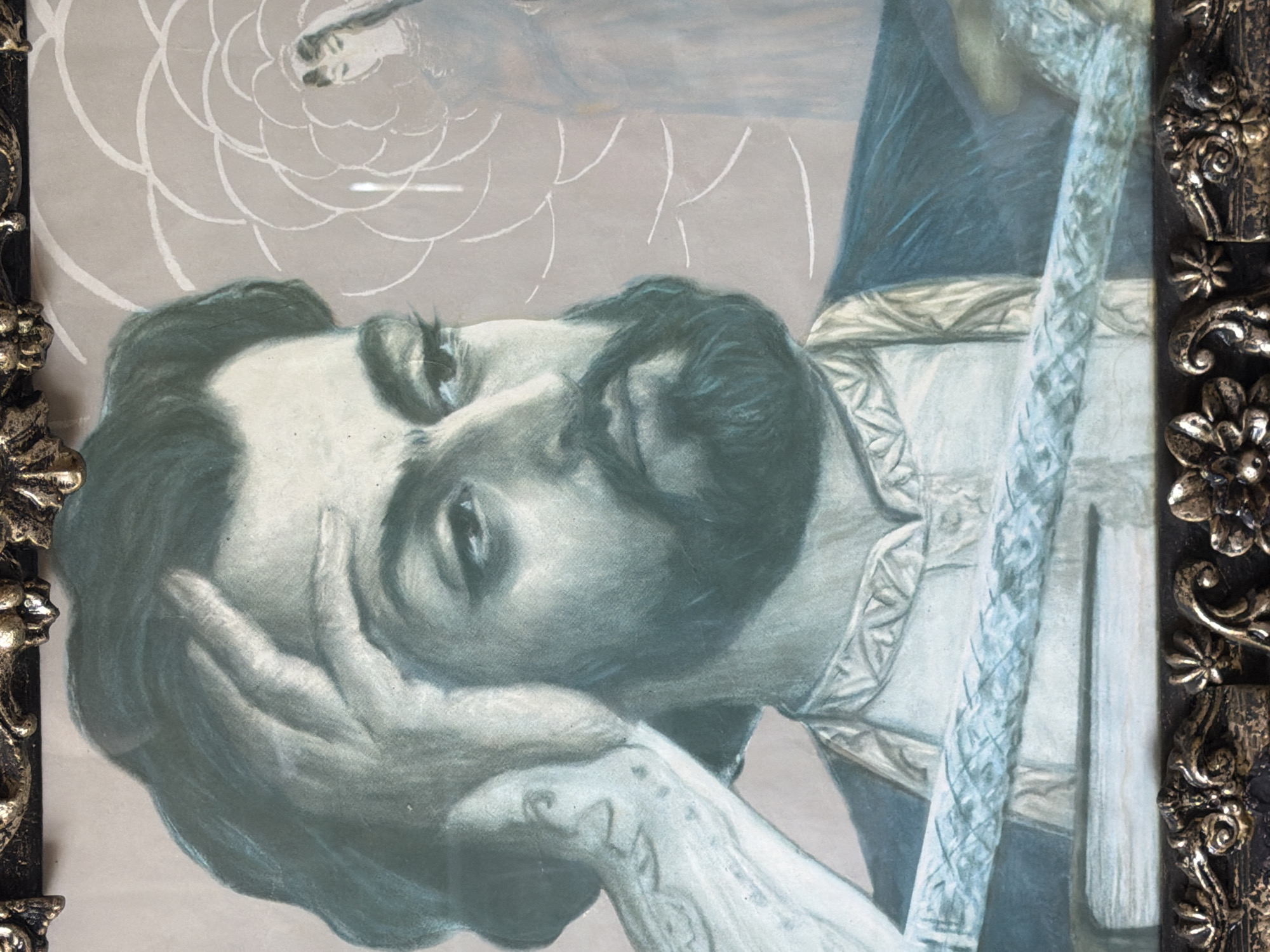 A framed portrait depicts a man reclining on his left side, his head resting on his left hand. The frame is ornate, with floral and scrollwork details in a gold-toned metal against a dark background. The man has dark hair and a beard, and he is looking upward with one eye visible. His shirt is light-colored with decorative trim. A braided rope-like object is positioned vertically in the upper right of the portrait. In the background, there is a faint image of a person superimposed over a white, floral-like pattern. The portrait is not indicative of a specific location.