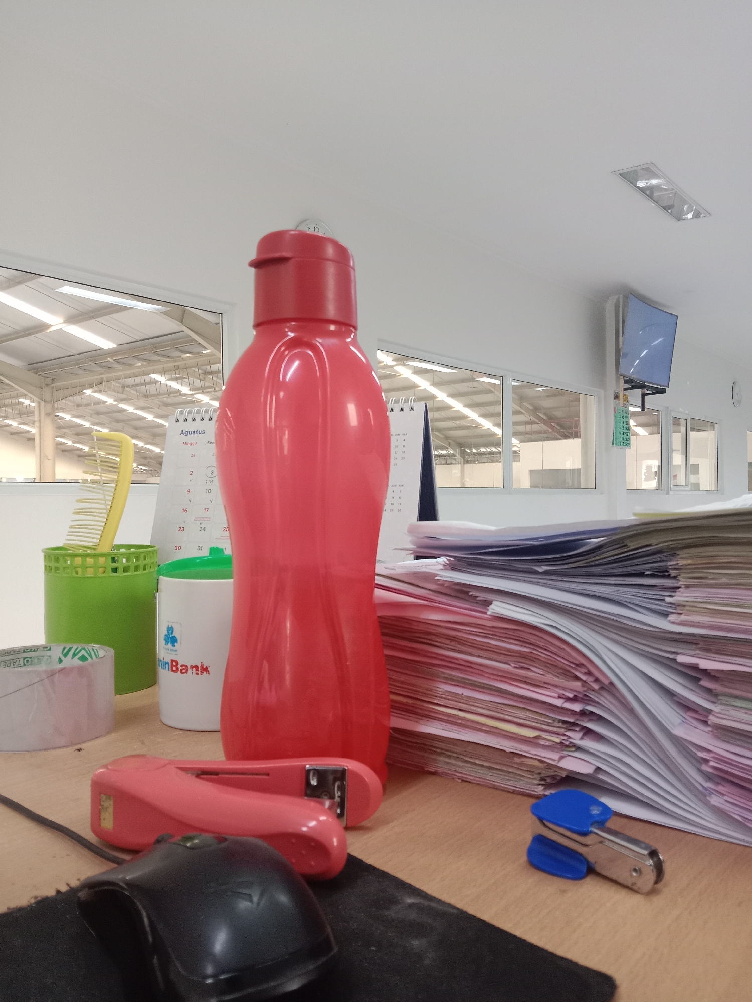 The image depicts a desk in an office environment. A large, red water bottle stands prominently in the center. To its left, a green container holds a yellow comb, and a roll of tape is partially visible. Next to the water bottle, a white cup with a logo and text that appears to be "Bank" is present. Behind these items, a desk calendar for August is visible, showing dates and days of the week. To the right of the water bottle, a large stack of papers is piled high. In the foreground, a pink stapler and a black computer mouse are positioned on the desk. The background shows large windows looking out onto an industrial or warehouse-like structure with a metal roof. The ceiling has fluorescent lighting fixtures. The overall impression is a functional workspace.