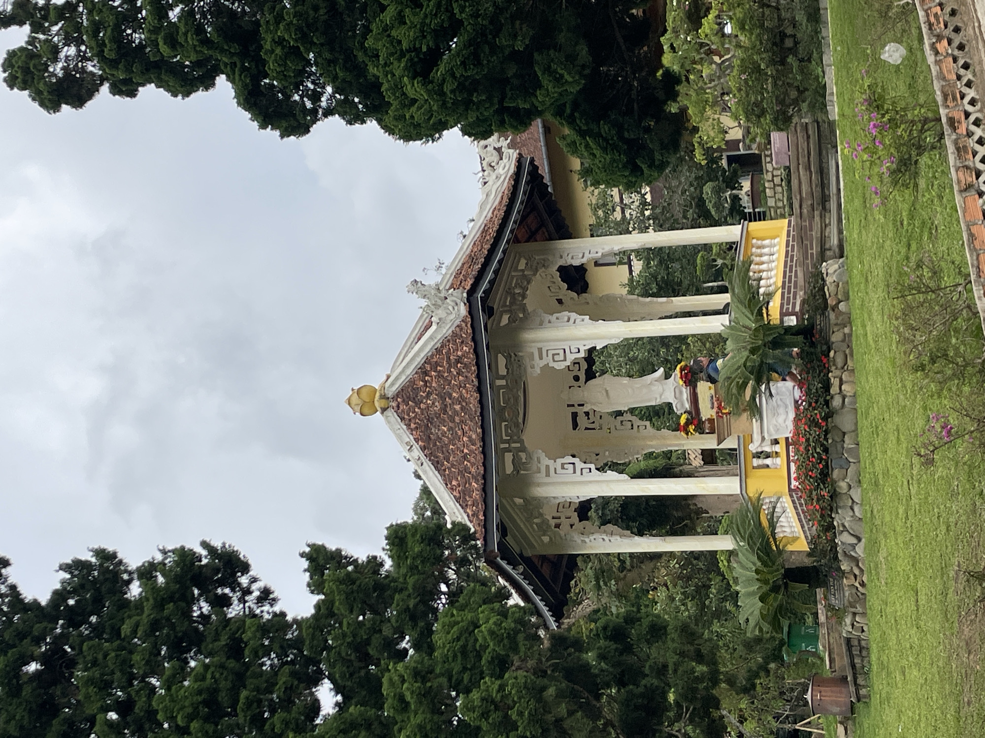 The image depicts the statue of a white figure, likely a deity or significant historical personage, housed within a decorative white pavilion. This pavilion features a traditional East Asian architectural style with a multi-tiered, terracotta-tiled roof adorned with decorative elements and a prominent golden finial. The pavilion is situated on a raised, yellow-painted platform, surrounded by a low, ornate white railing.
In front of the pavilion, a vibrant display of red flowers and lush green foliage creates a colorful foreground. To the left of the pavilion, a tall, dark green evergreen tree frames the structure, with its dense foliage creating a dramatic contrast against the overcast sky. On the right, another similar evergreen tree and various smaller green bushes and trees contribute to the lush, verdant environment.
The pavilion itself is supported by white pillars and features intricate latticework and decorative carvings on its exterior. Behind the pavilion, a building with a yellow facade is visible, partially obscured by the trees. The ground in front of the pavilion is a well-maintained green lawn, with a low stone wall and more flowering plants adding to the aesthetic appeal of the landscape.
This scene is located at the Quan The Am Pagoda in Da Lat, Vietnam. The pagoda is known for its serene atmosphere and beautiful gardens, making it a popular destination for both religious pilgrimage and tourism. The overcast sky suggests a possibly humid or rainy day, common in Da Lat's tropical highland climate. The overall impression is one of tranquility and natural beauty, with the pavilion serving as a focal point within the meticulously landscaped surroundings.