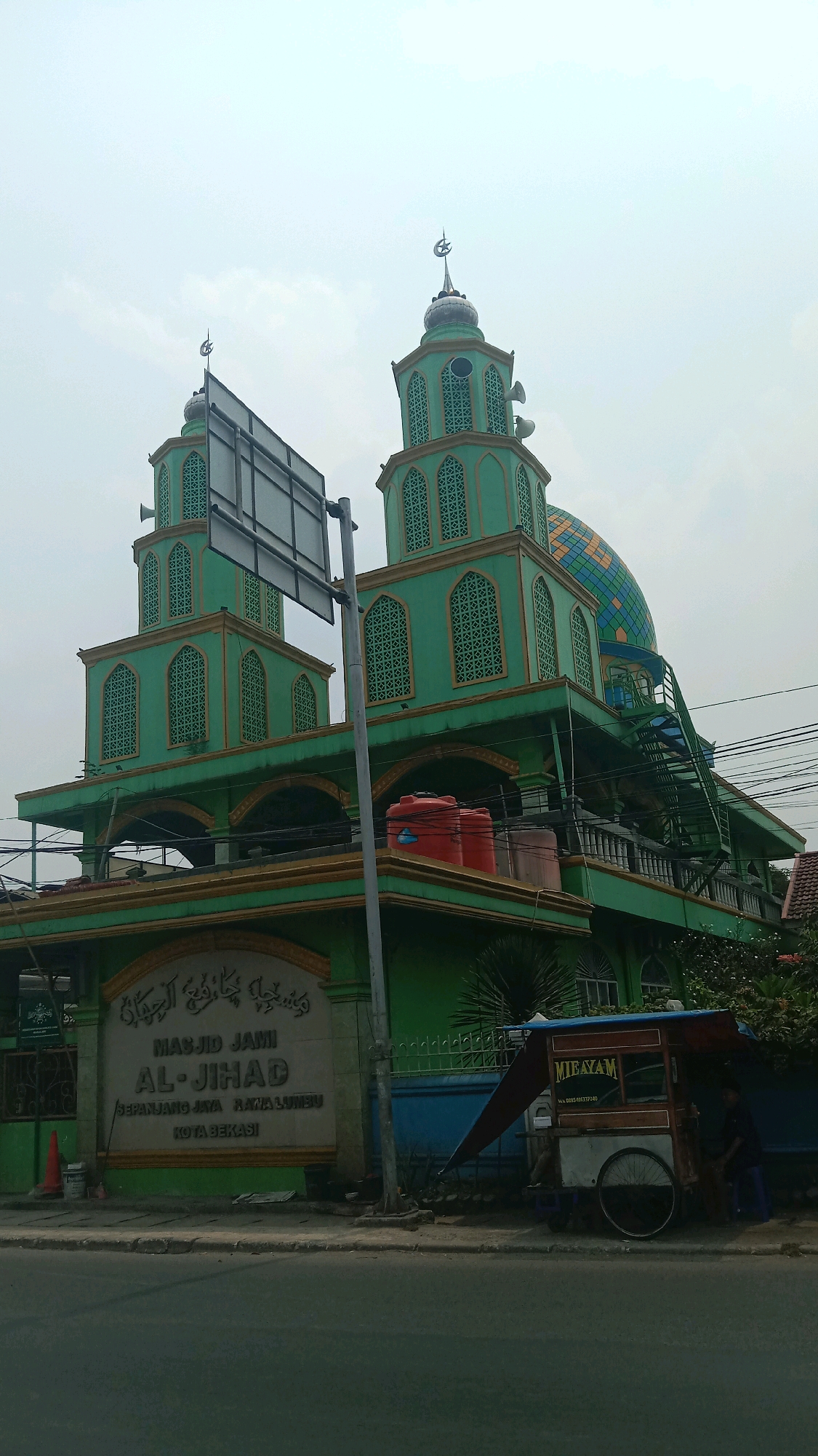 An exterior, eye-level shot captures the Masjid Jami Al-Jihad in Kota Bekasi, Indonesia, under a hazy, overcast sky. The mosque's dominant feature is its vibrant green facade, accented with gold trim. Two prominent minarets, also green with gold detailing and topped with crescent moons, rise on either side of a large, colorful dome. The dome itself is a mosaic of blue and yellow geometric patterns.
Below the main structure, a building with arched openings and balconies is visible, with two large red water tanks situated on one of the balconies. A sign with Arabic script and the mosque's name, "MASJID JAMI AL-JIHAD," along with its address "SEPANJANG JAYA RAJA LUMBU KOTA BEKASI," is prominently displayed at the entrance.
To the right of the mosque, a mobile food cart with a blue tarp and "MIE AYAM" signage is parked on the sidewalk. A person is visible behind the cart. A large, dark sign stands near the left minaret. The scene is bordered by a paved road in the foreground and some greenery and trees to the right, suggesting a location within a developed urban area. Electrical wires are visible strung across the scene.