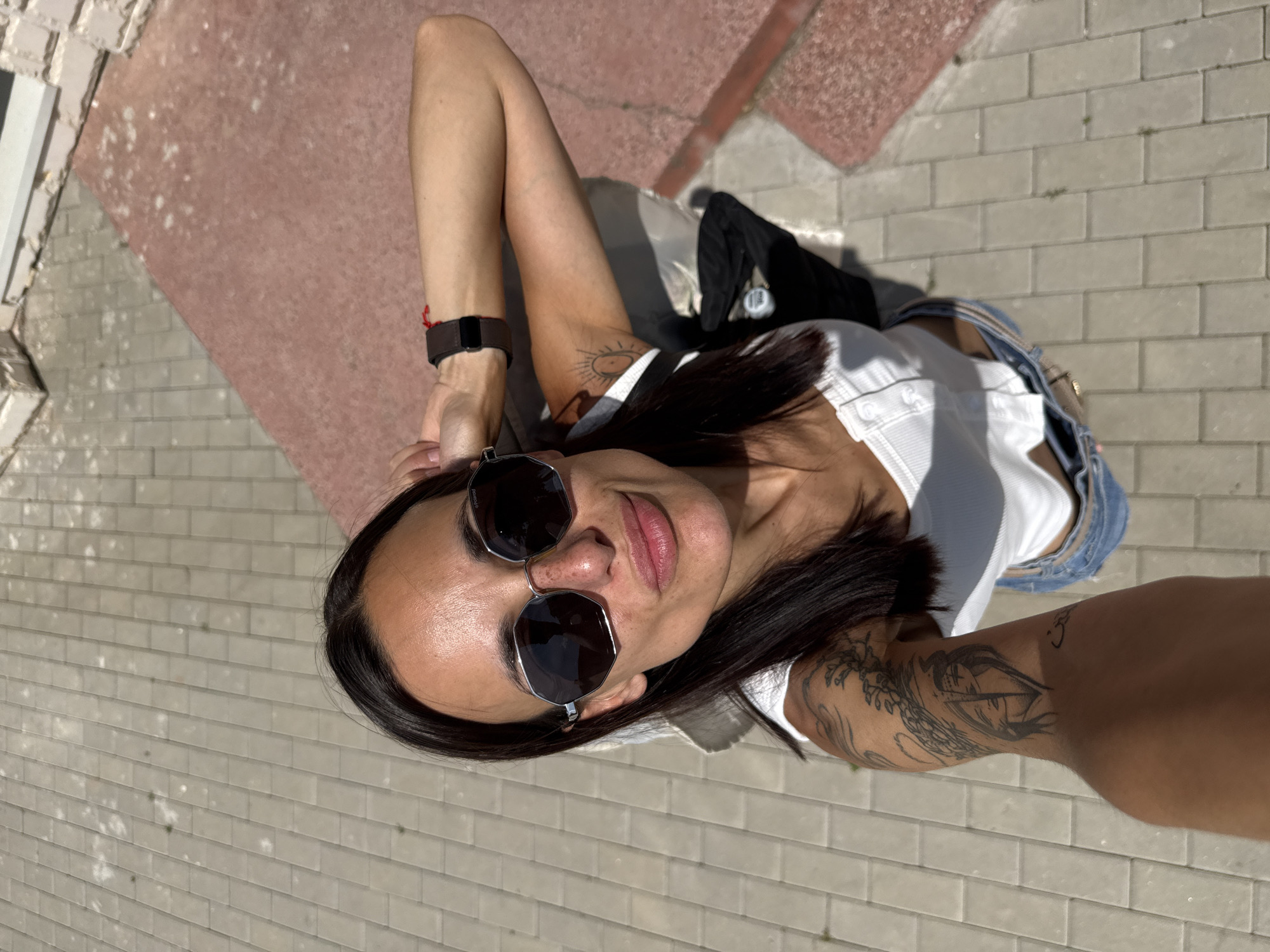 A woman is taking a selfie outdoors. She is wearing sunglasses, a white top, and denim shorts. Her arm is resting on her head, and she has tattoos on her arm. The background shows a paved walkway and a pinkish-red area. The scene appears to be in Pordim, Bulgaria.