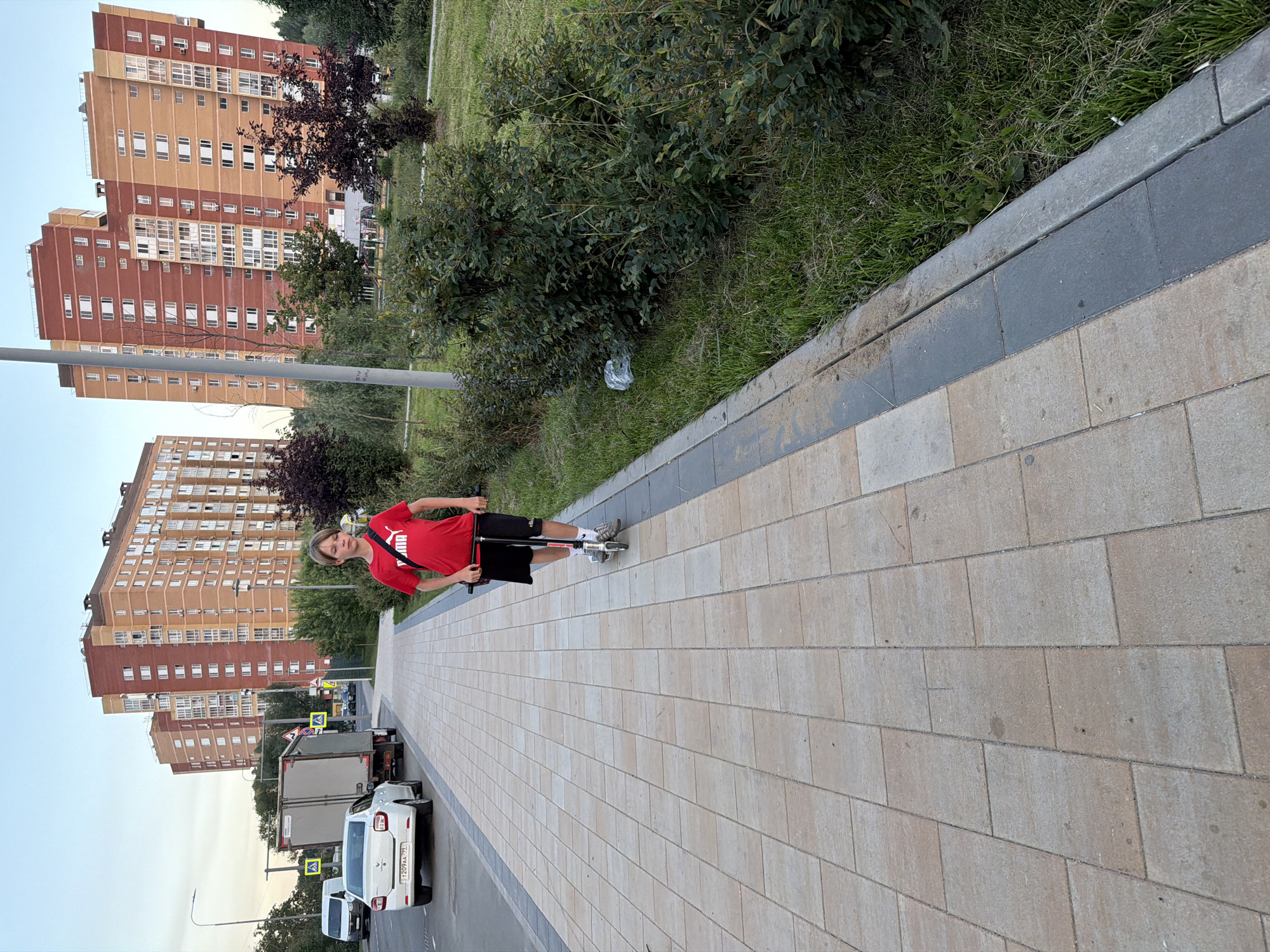 A boy rides a scooter on a paved sidewalk in the city of Shcherbinka, Russia. He is wearing a red shirt and black shorts. The sidewalk is lined with grass and bushes. In the background, there are several tall residential buildings and a truck. The sky is clear and blue.