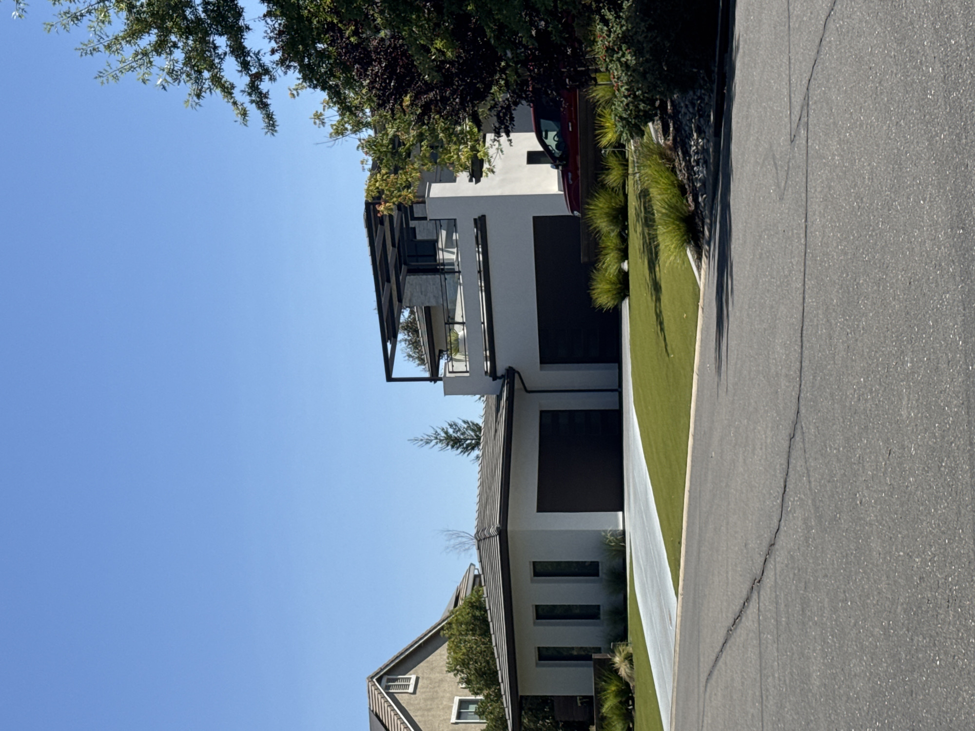 The image shows a modern, multi-level white stucco house with a flat roof, located in Roseville, United States. The house features a two-car garage with dark garage doors, and a second level with a large balcony or patio area covered by a modern black pergola. A red car is parked in front of the house. The property is landscaped with green grass, bushes, and some rocks. The scene is captured on a clear, sunny day with a bright blue sky. The foreground shows a cracked asphalt road. To the left of the main house, a portion of another house with a sloped, tiled roof and solar panels is visible.