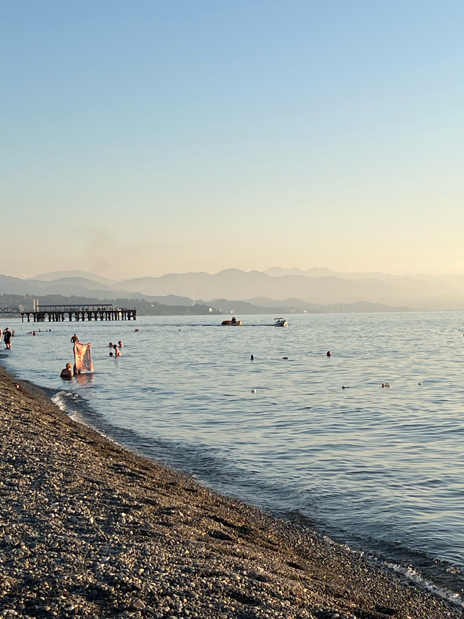 The image depicts a tranquil scene at a pebble beach in Kobuleti, Georgia, during what appears to be late afternoon or early evening. The sea is calm with gentle ripples, reflecting the soft hues of the sky. In the foreground, a dense layer of various sized grey pebbles lines the shore, with small waves lapping at the edge.
The water is populated by several individuals. Some are wading near the shore, while others are swimming further out. One person is holding up a colorful fabric, possibly a towel or sarong, near the water's edge. In the middle ground, a pier extends into the sea, with people visible on it and around it.
Further out in the sea, two small boats are present. One is a speed boat, cutting through the water, and the other appears to be a smaller vessel. Scattered across the water are several buoys, some red and some white.
The background features a series of hazy, layered mountains that fade into the soft, pale blue sky, suggesting a clear but slightly hazy atmosphere. The overall impression is one of a relaxed summer day at a coastal resort.