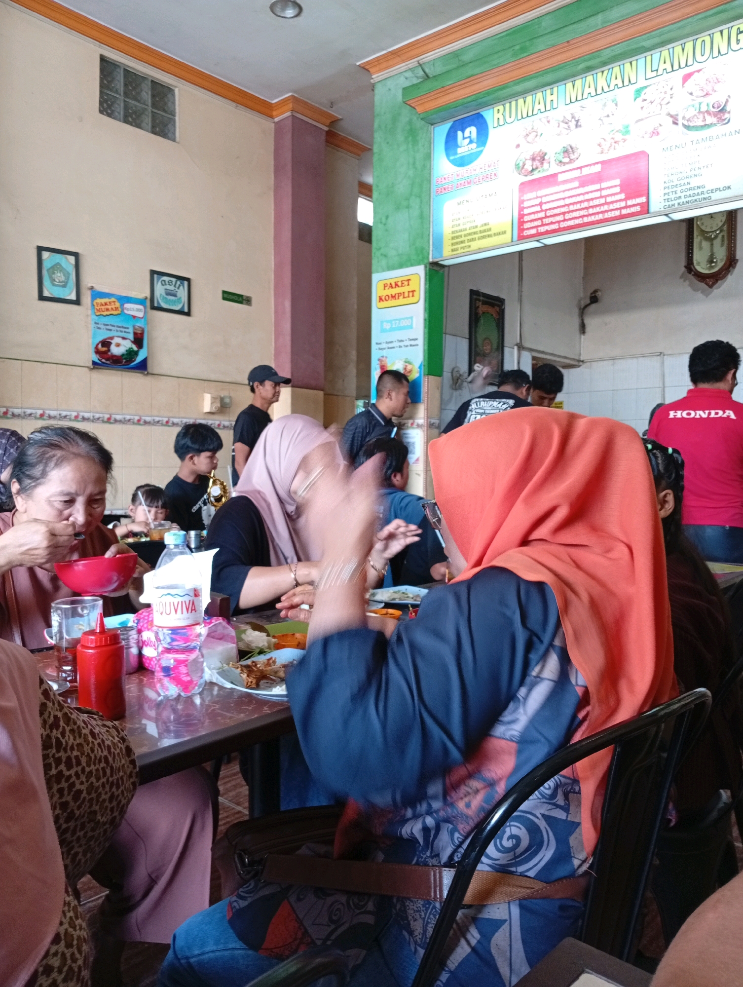 The image depicts the interior of a casual dining establishment, identified by the signage "RUMAH MAKAN LAMONG" in the upper right portion. The setting appears to be a local eatery in Kongsijaya, Indonesia, as indicated by the presence of Indonesian language menus and the general ambiance. The scene is populated by several individuals seated around tables, engaged in eating and socializing.
In the foreground, a woman on the left is eating from a red bowl with a spoon, while another woman on the right, partially obscured by an orange hijab, is also seated at the table. A bottle of "AQUVIVA" water and a red sauce dispenser are visible on the table. In the background, other patrons and staff members are present. A man in a black t-shirt is visible to the left, and a person wearing a red shirt with "HONDA" printed on it is on the far right.
The restaurant interior features light-colored walls, a tiled floor, and overhead lighting. Several posters and signs are displayed on the walls, including menu boards and promotional materials. The overall atmosphere suggests a busy, informal dining environment.