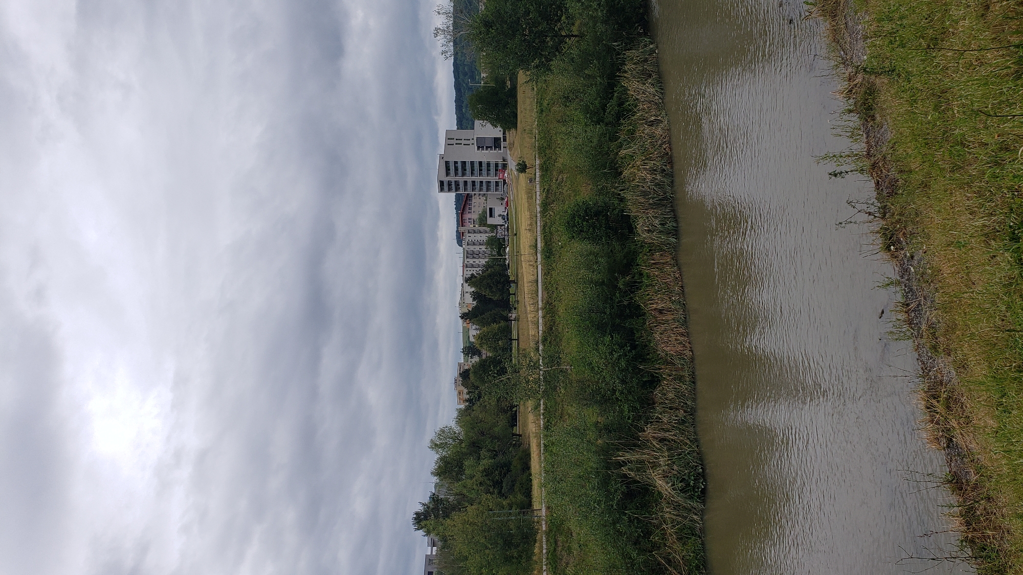 The image shows a cloudy day scene in Prešov, Slovakia. In the foreground, a muddy river flows, with grassy banks and some reeds along the edges. Across the river, a green field stretches out, bordered by trees and then a cityscape. The cityscape features several multi-story apartment buildings and a prominent modern, white building with multiple balconies. The sky is overcast with gray clouds, indicating an impending or recent rain. The overall impression is a moderately urban landscape with elements of nature.