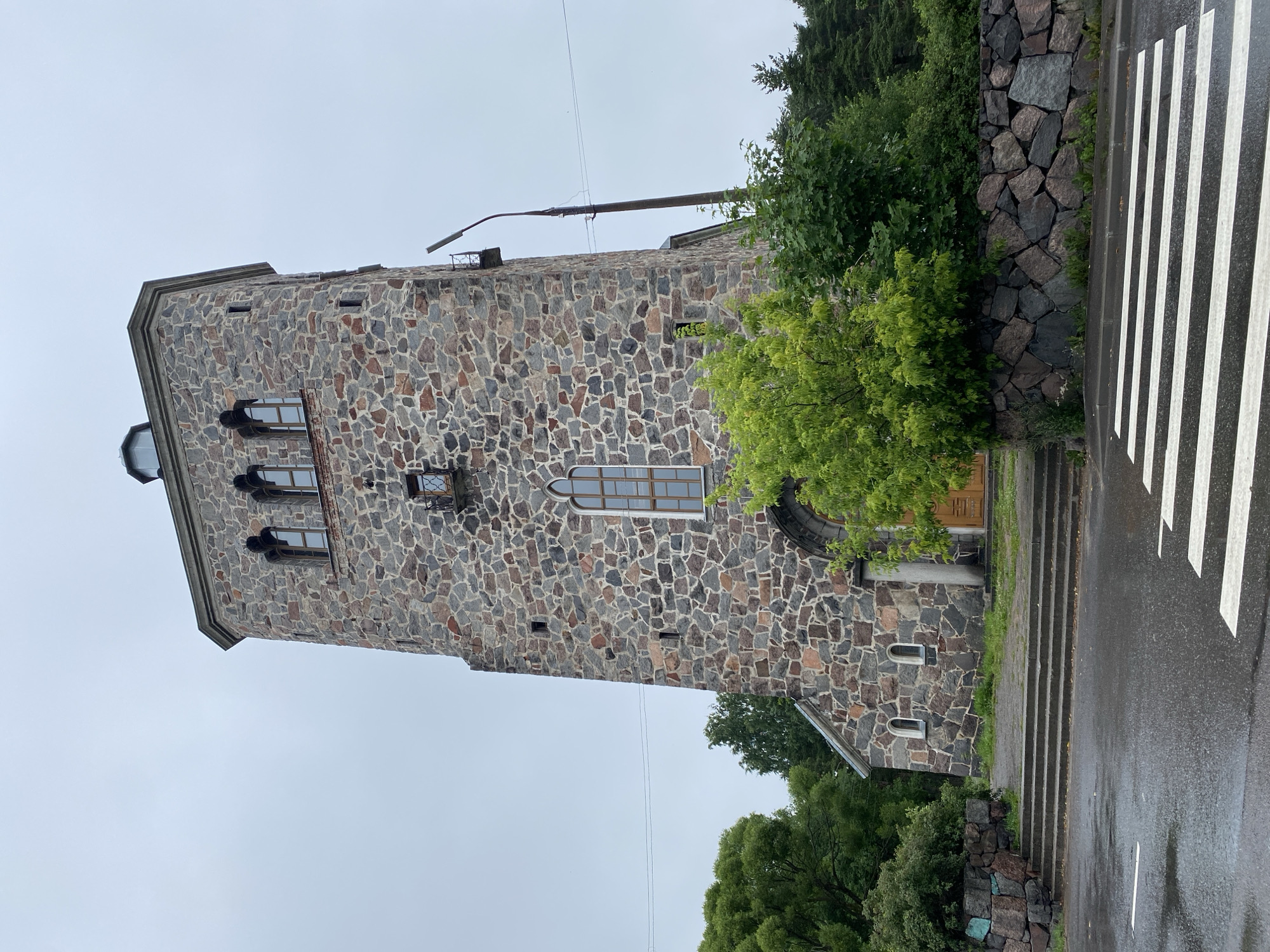 The image shows a stone tower, likely a church or bell tower, located in the city of Priozersk, Russia. The tower is constructed from rough-hewn, multi-colored stones, giving it a rustic and sturdy appearance. It has a square base that transitions to an octagonal top. Several windows are visible, some with arched tops and wooden frames. A small balcony or ledge projects from one side of the tower. The tower is surrounded by lush green trees and vegetation. In the foreground, a wet, paved road with a white crosswalk is visible, indicating a street or pedestrian crossing. The sky is overcast, suggesting a cloudy or possibly rainy day. The overall impression is of an old, historic structure set within a natural, green environment.