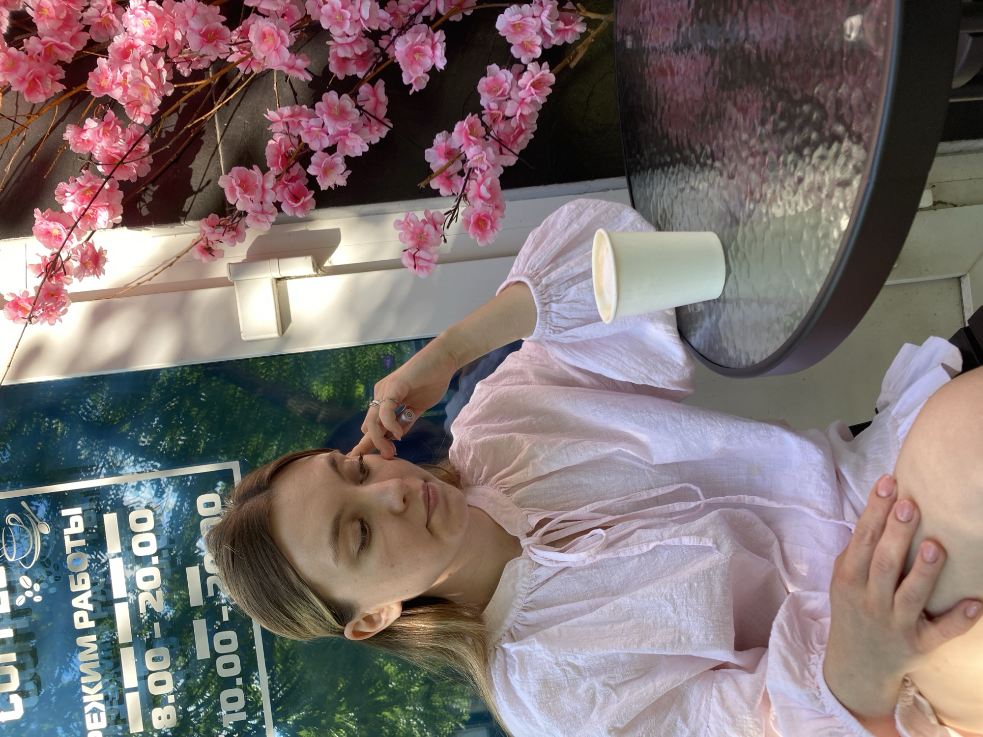 In Chaikovsky, Russia, a young woman sits at a glass-topped table outside a cafe. She is wearing a light pink, crinkled blouse with voluminous sleeves and a tie at the neckline. Her hand rests on her thigh, and she is looking down with her fingers lightly touching her temple. A white paper cup of coffee sits on the table to her right. Behind her, a window displays the cafe's operating hours: "РЕЖИМ РАБОТЫ 8.00 - 20.00 10.00 - 20.00." To the right of the woman, artificial pink cherry blossoms adorn branches against a dark wall. Sunlight casts shadows on the scene, suggesting it is daytime.