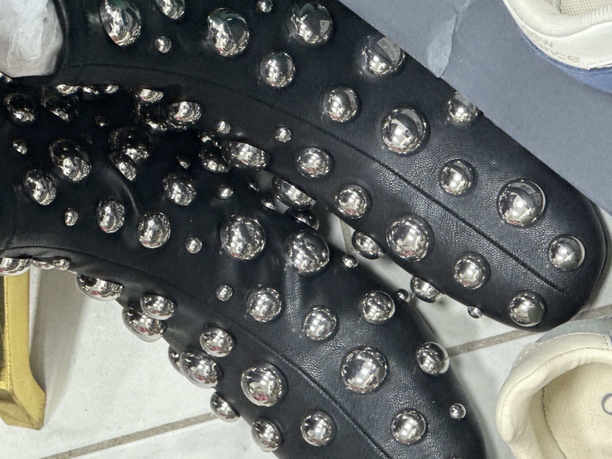 The image shows a pair of black, studded boots on a tiled floor, adjacent to a white sneaker and a golden object. The boots are decorated with numerous shiny, silver-colored studs of varying sizes. The overall environment appears indoors. There is no evidence that the scene is located in Chaikovsky, Russia.