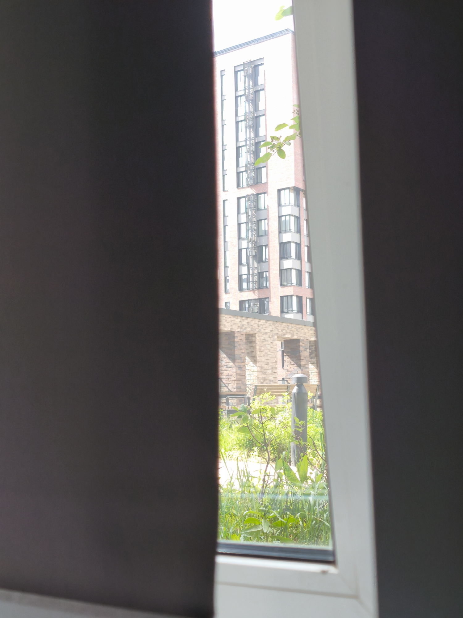 A partially obscured view through a window shows an exterior scene of a building facade and courtyard in Moscow, Russia. The window frame is visible, with a dark curtain partially obstructing the view. The scene outside includes a multi-story building with a fire escape, a brick structure, and a courtyard area with greenery. A tall, gray lamp post stands among the plants.